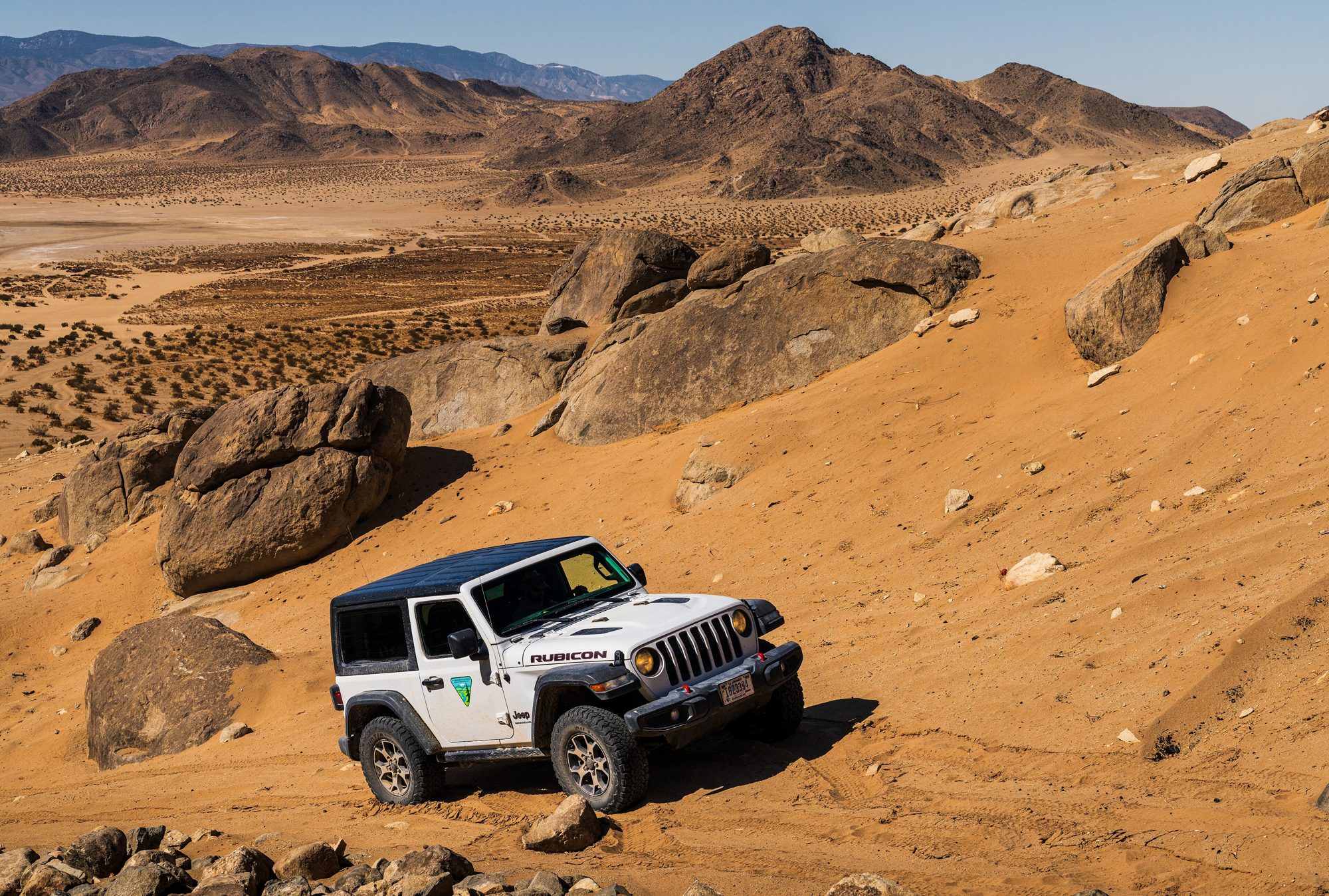 A white two-door Jeep Wrangler JL Rubicon climbing a sandy slope between granite boulders