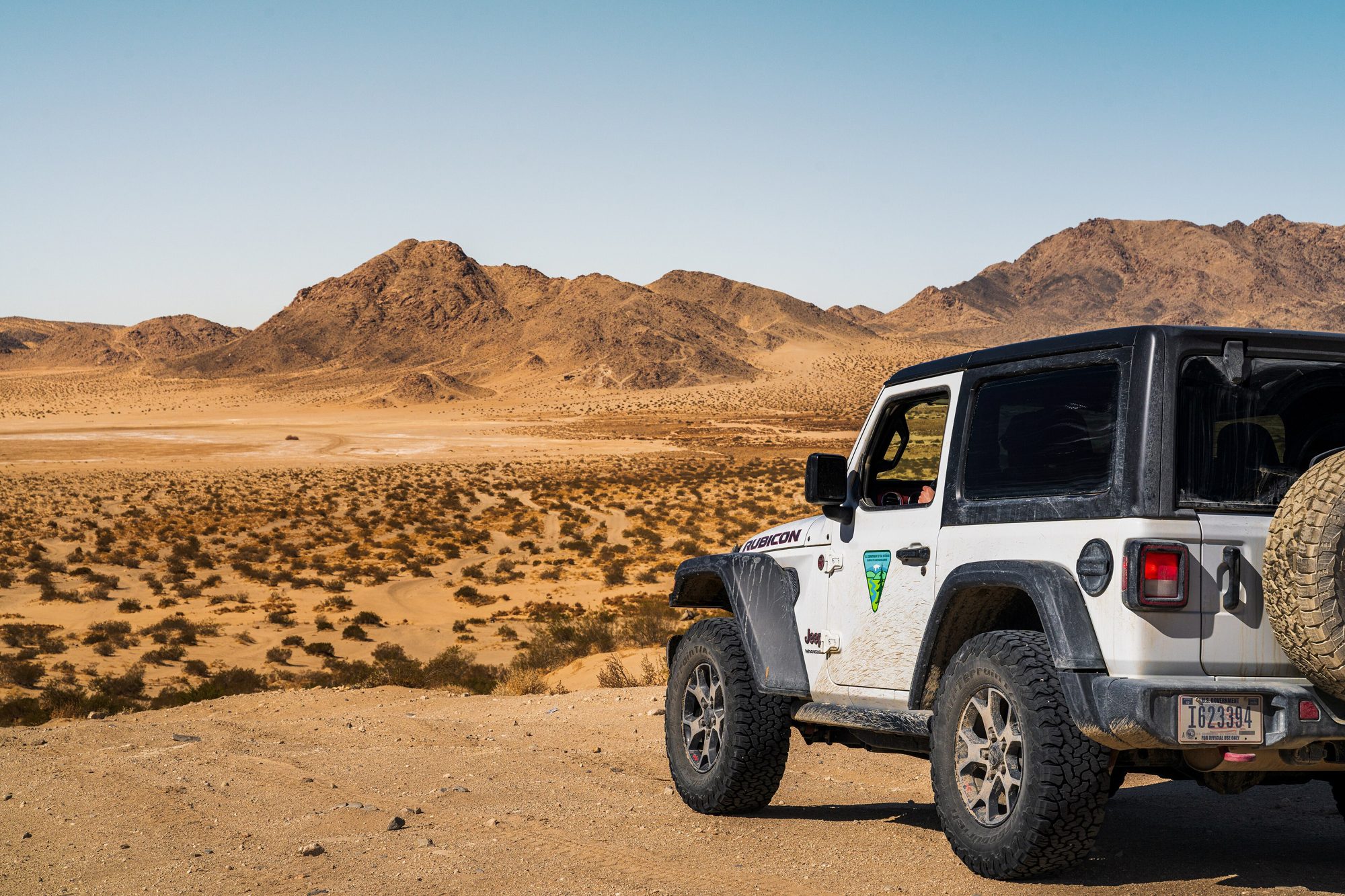 Rear three-quarter view of a white two-door Jeep Wrangler JL with its spare wheel, parked above a desert valley