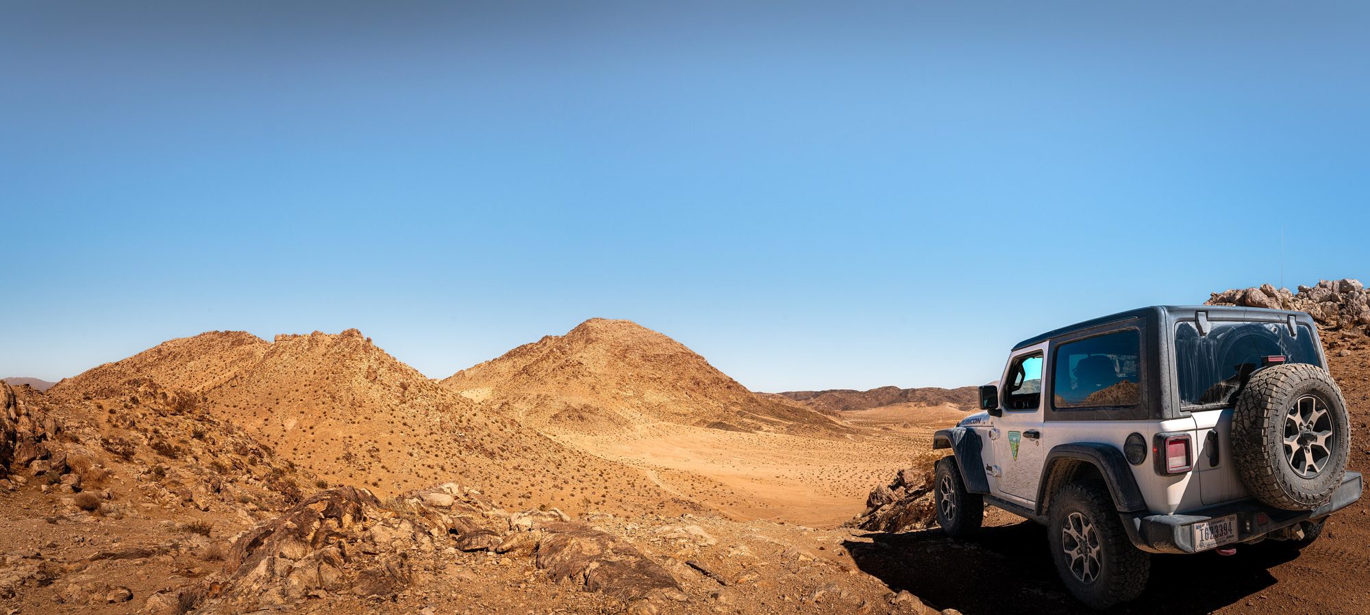 A white two-door Jeep Wrangler JL Rubicon on a rocky desert trail with brown mountains behind
