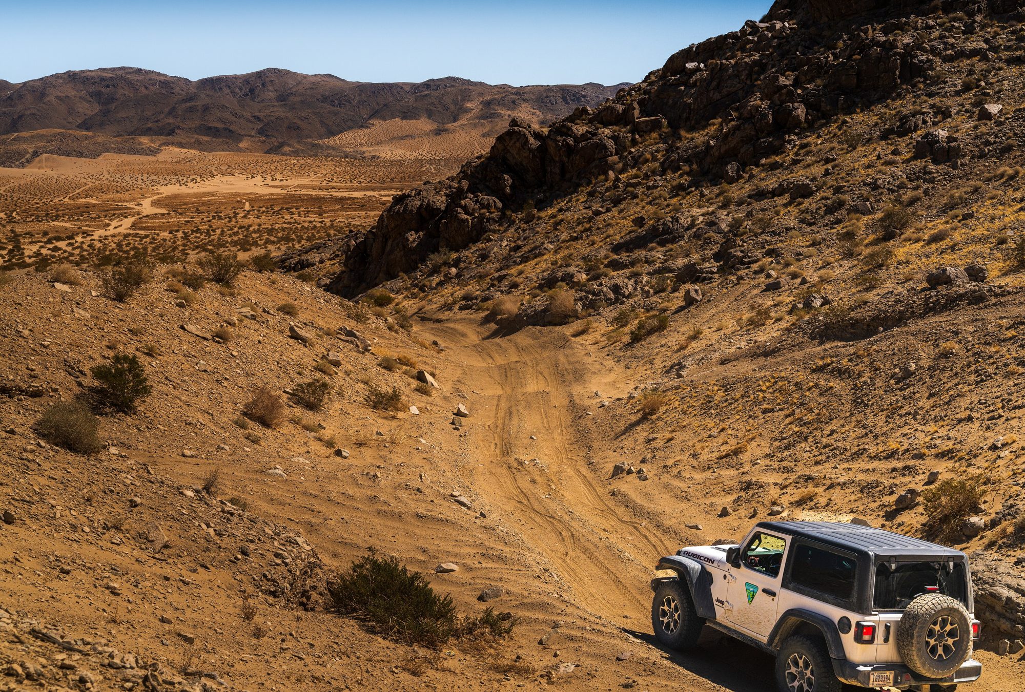A white two-door Jeep Wrangler JL, seen from behind, on a sandy wash trail below rocky desert hills