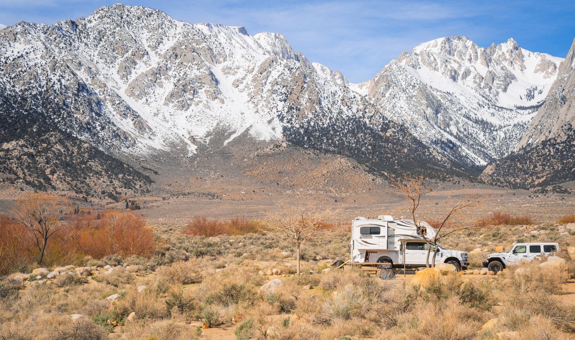 A white Jeep Wrangler JL Unlimited at a campsite beside a truck camper below the snow-capped Sierra Nevada