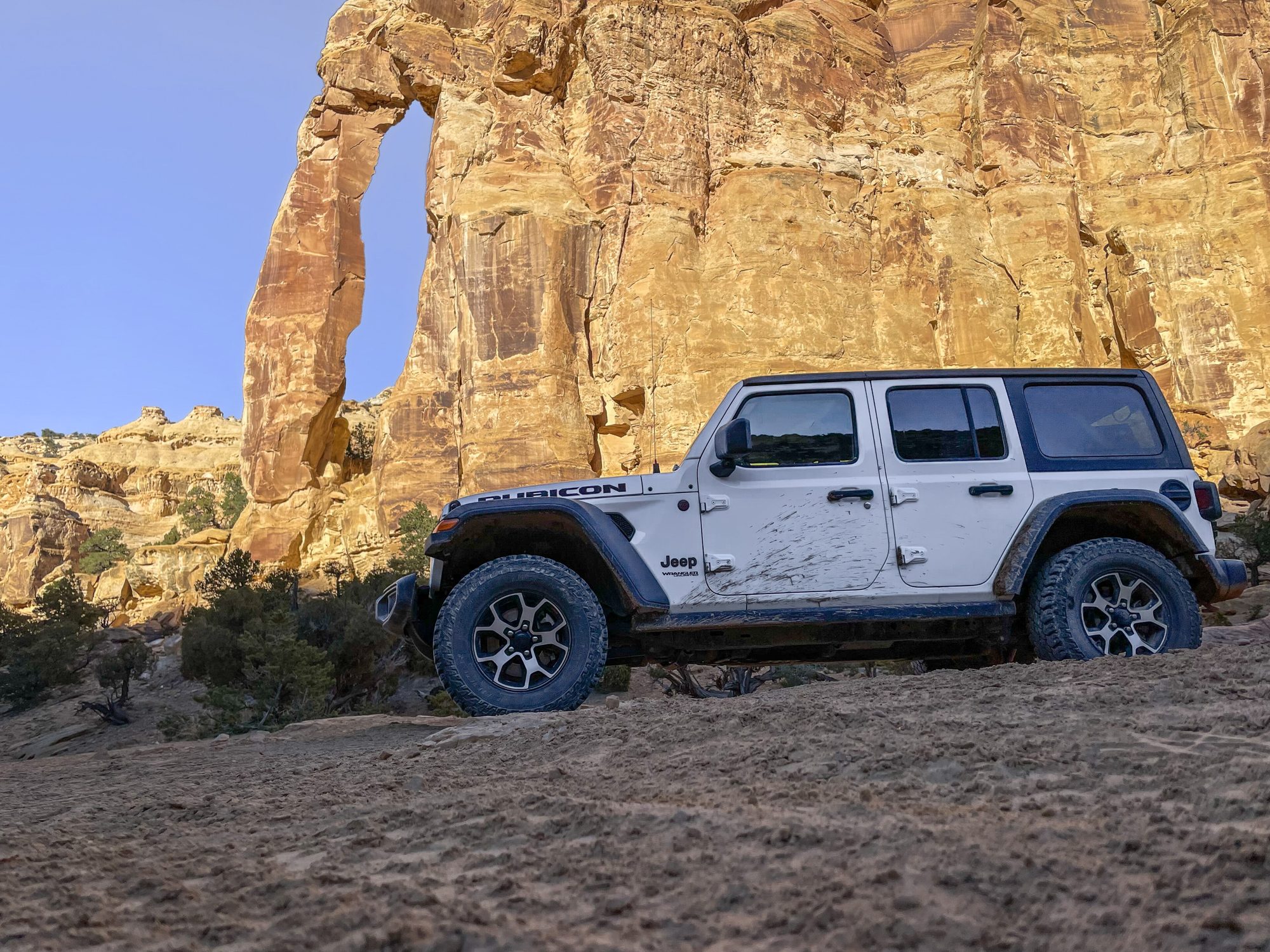 A white Jeep Wrangler JL Unlimited parked on a dirt track beneath Eagle Arch, a sandstone arch in Utah's San Rafael Swell