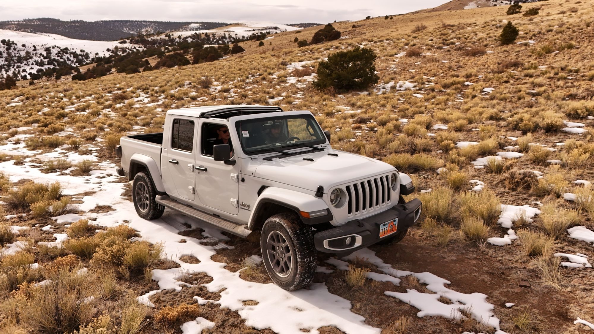 A white pre-2024 Jeep Gladiator Overland with its roof panels off, driving a snowy two-track through sagebrush hills