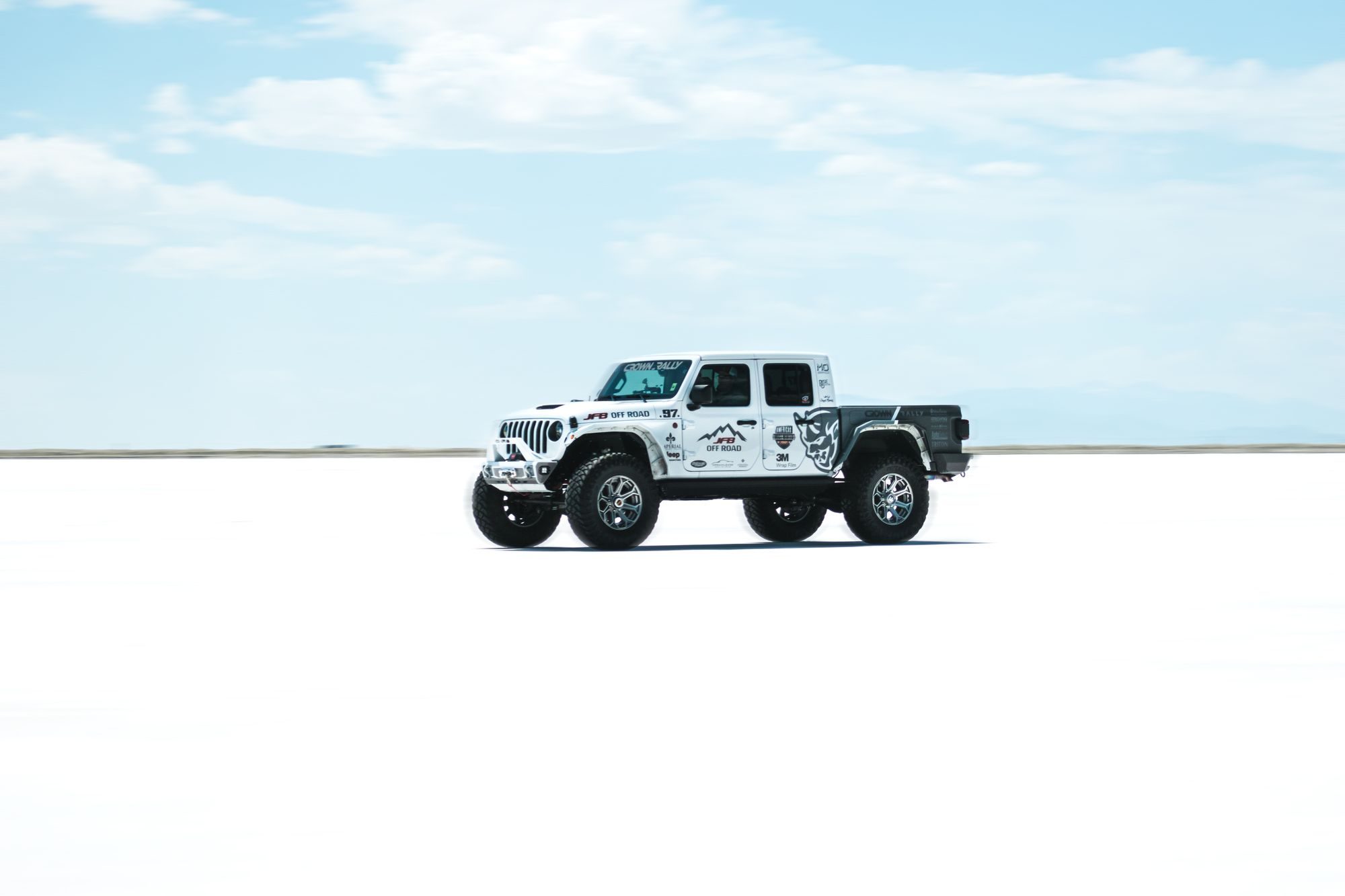 A heavily modified white rally-liveried Jeep Gladiator on big tires crossing a salt flat under a blue sky