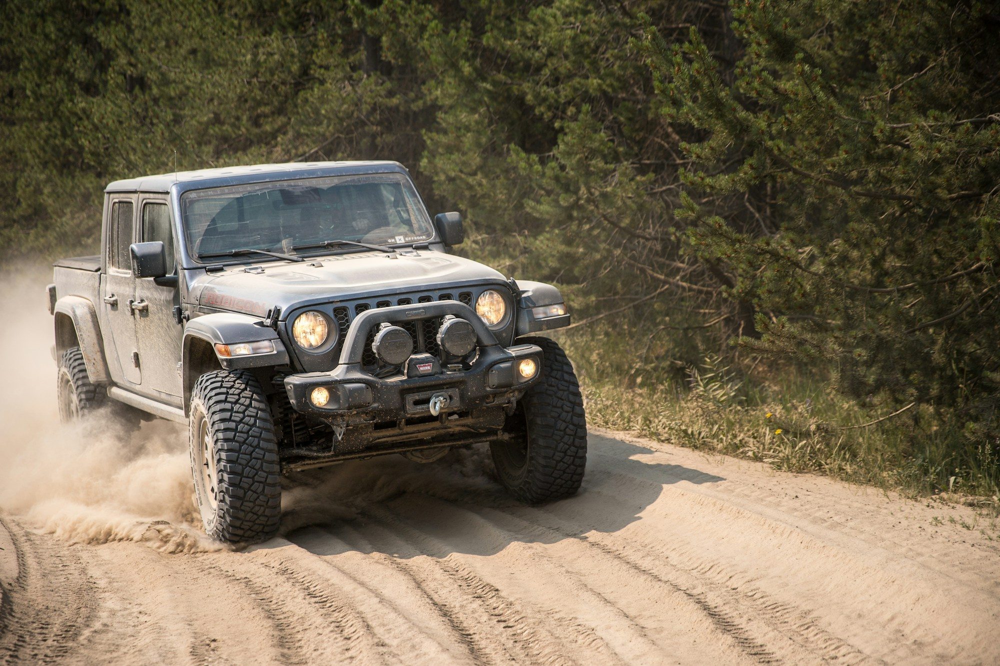 A grey Jeep Gladiator Rubicon with a steel winch bumper and auxiliary lights kicking up dust on a sandy forest road