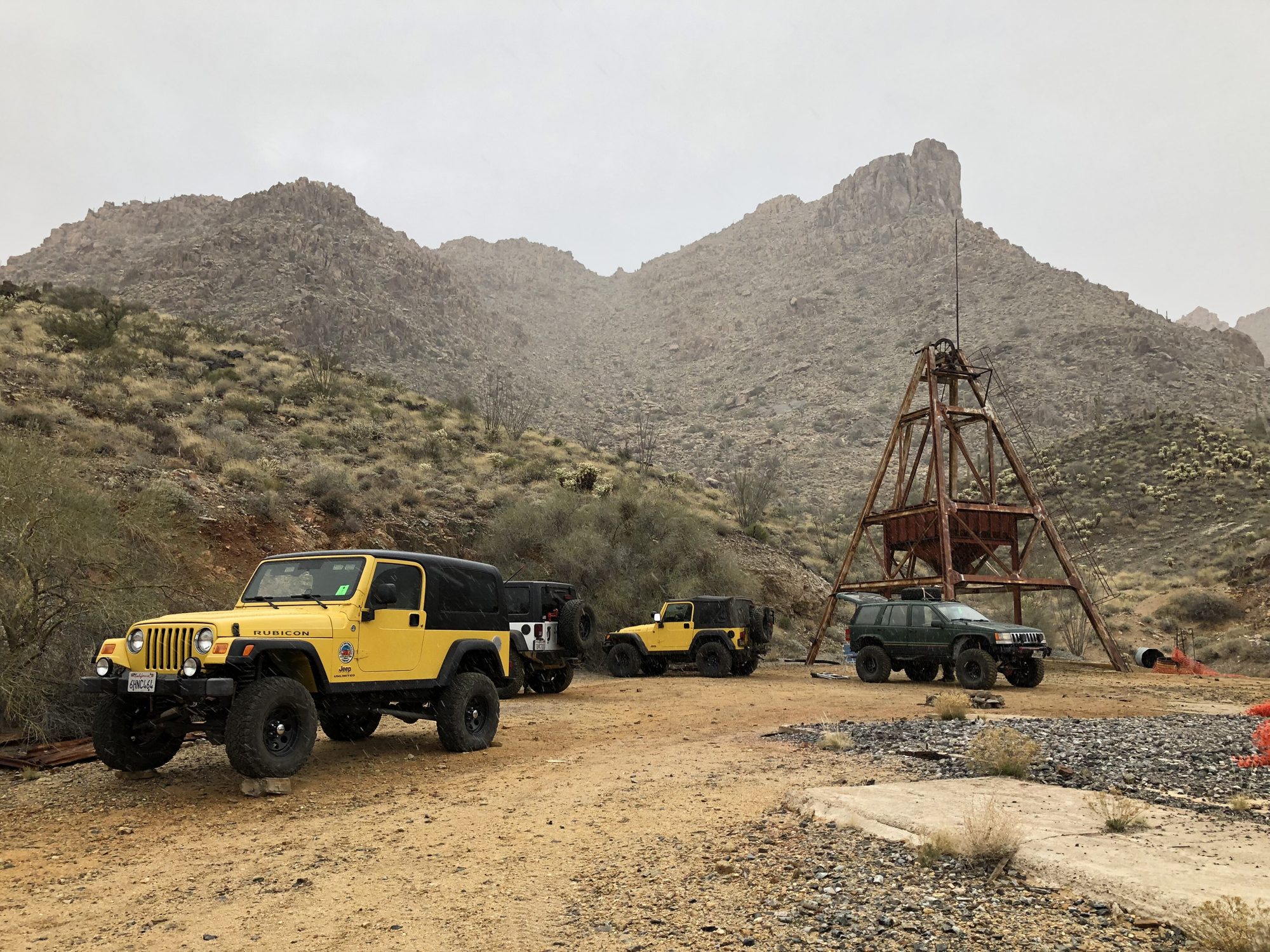 A yellow Wrangler Unlimited Rubicon (LJ) with a black hardtop parked on a dirt track in desert mountains beside an old mine headframe, with more Jeeps behind