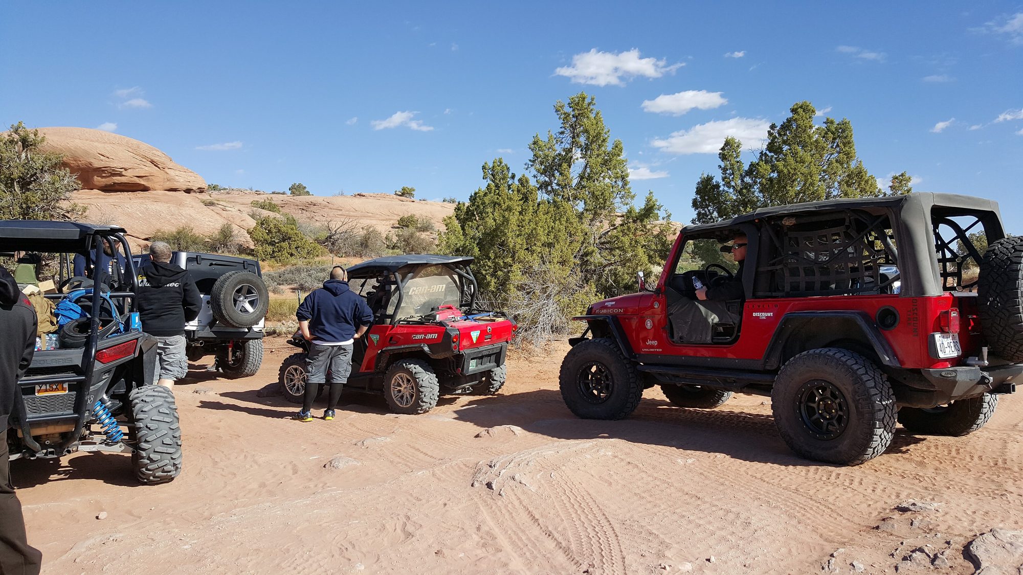 A red lifted Wrangler Unlimited Rubicon (LJ) with a roll cage on red sand beside juniper scrub on the Hell's Revenge trail near Moab