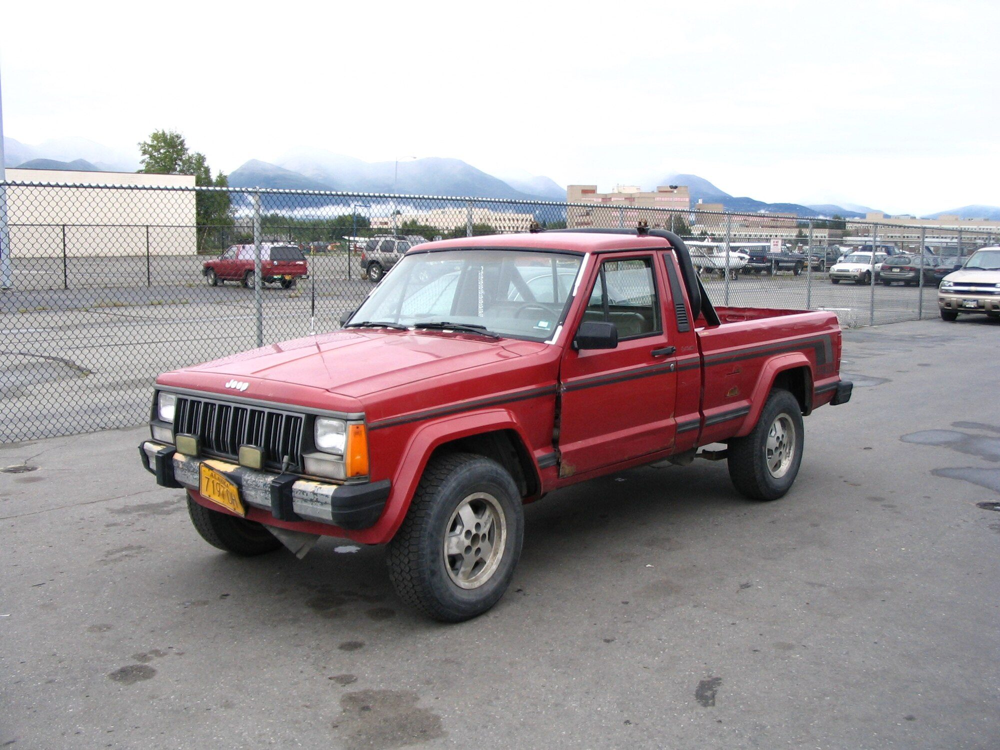 A red short-bed Jeep Comanche with a roll bar and auxiliary lights in a lot below cloud-capped mountains