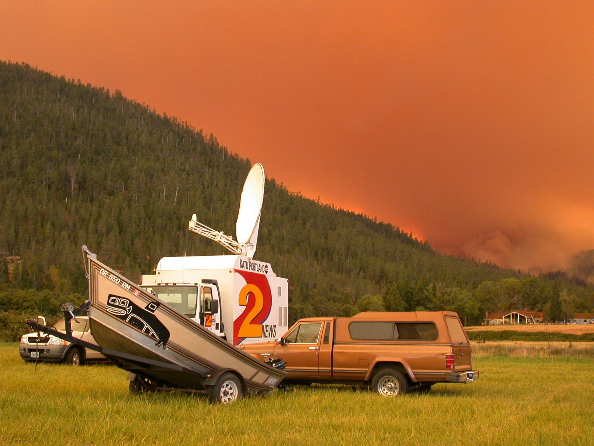 A tan long-bed Jeep Comanche with a camper shell parked in a grassy meadow below a forested mountain, with orange wildfire smoke filling the sky