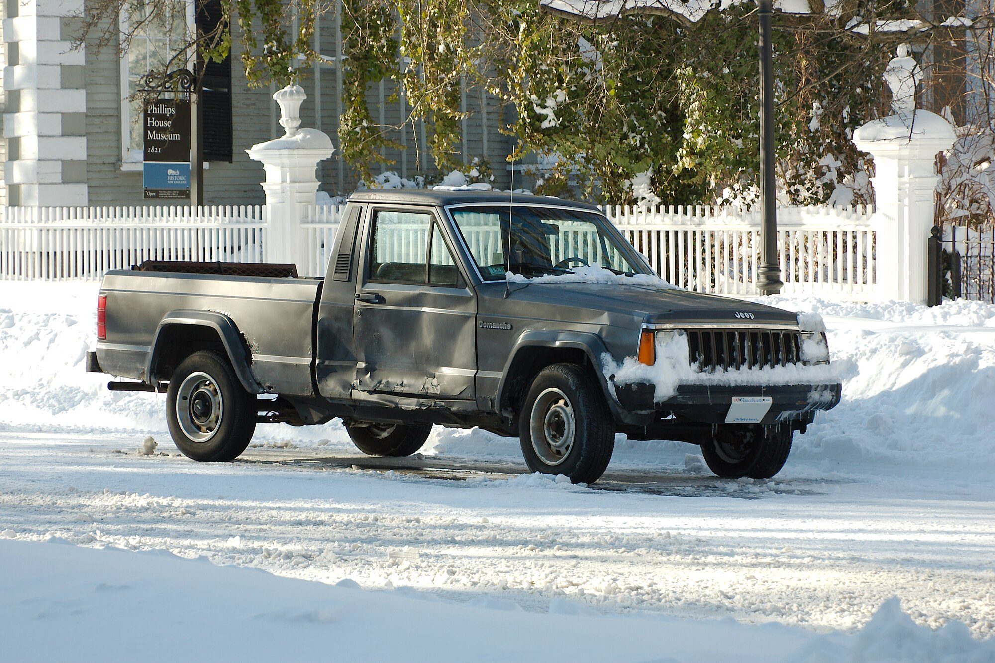 A weathered grey long-bed Jeep Comanche parked in deep snow on a plowed street