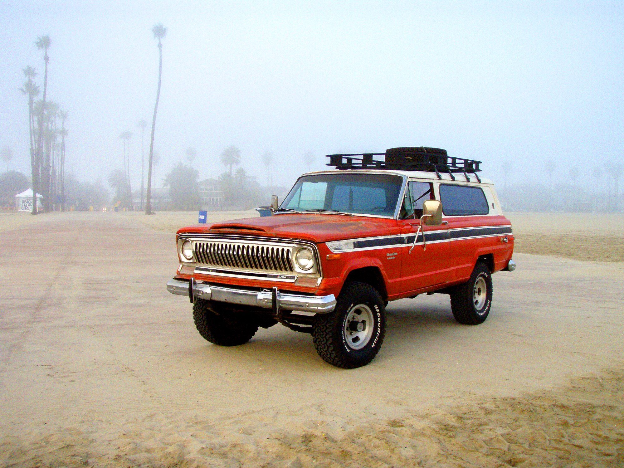 A red and white 1974 Jeep Cherokee S on BFGoodrich all-terrains with a roof basket and spare, parked on a foggy sand beach lined with palm trees