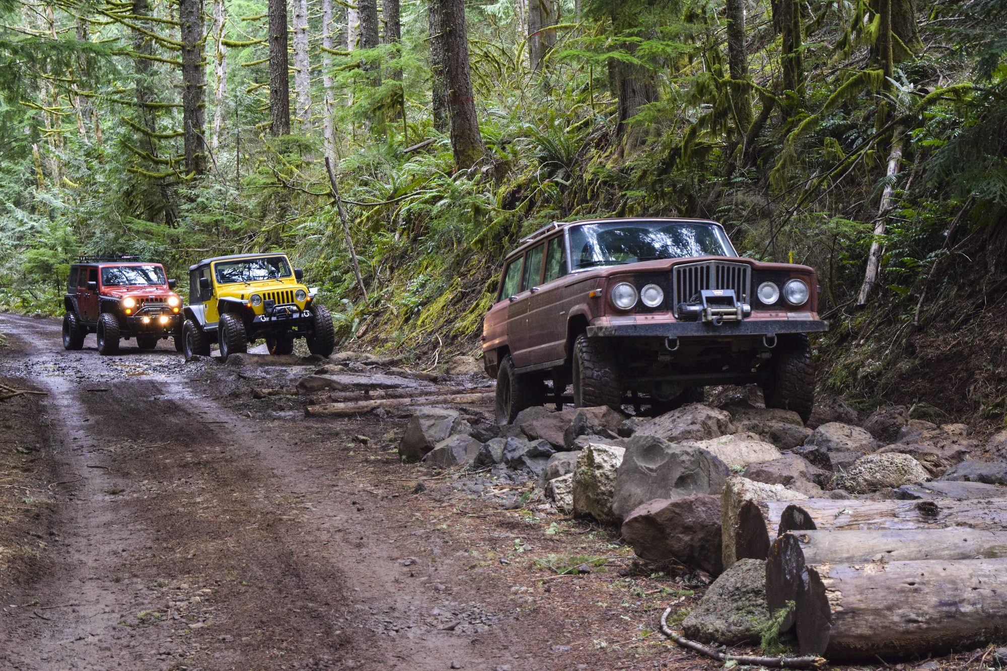 A lifted, faded red early SJ Wagoneer with a winch bumper climbing over basalt boulders on a muddy forest trail, with two Wranglers behind