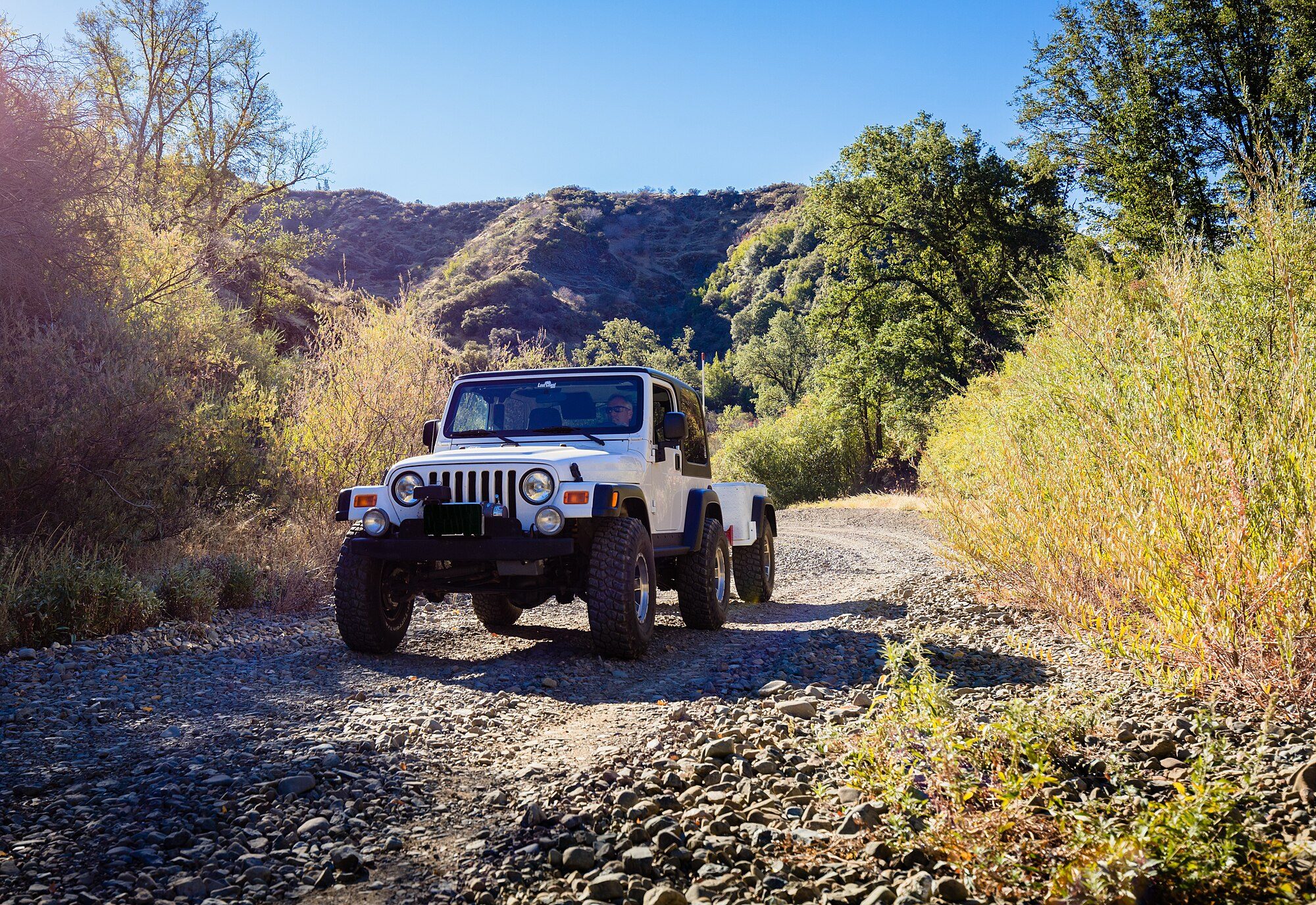 A lifted white Jeep Wrangler TJ towing a small off-road trailer on a gravel trail through autumn brush at South Cow Mountain