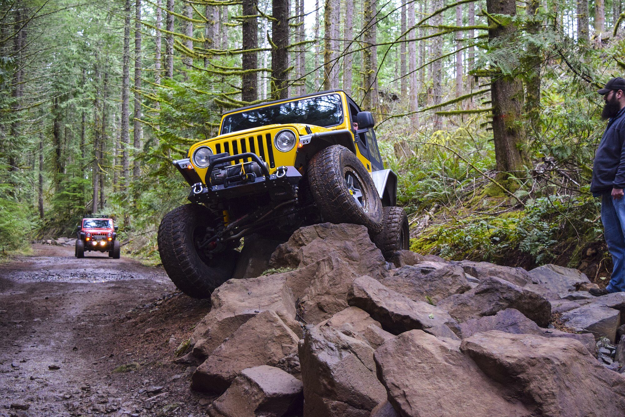 A lifted yellow Jeep Wrangler TJ climbing over a pile of rocks on a forest trail, with a spotter beside it