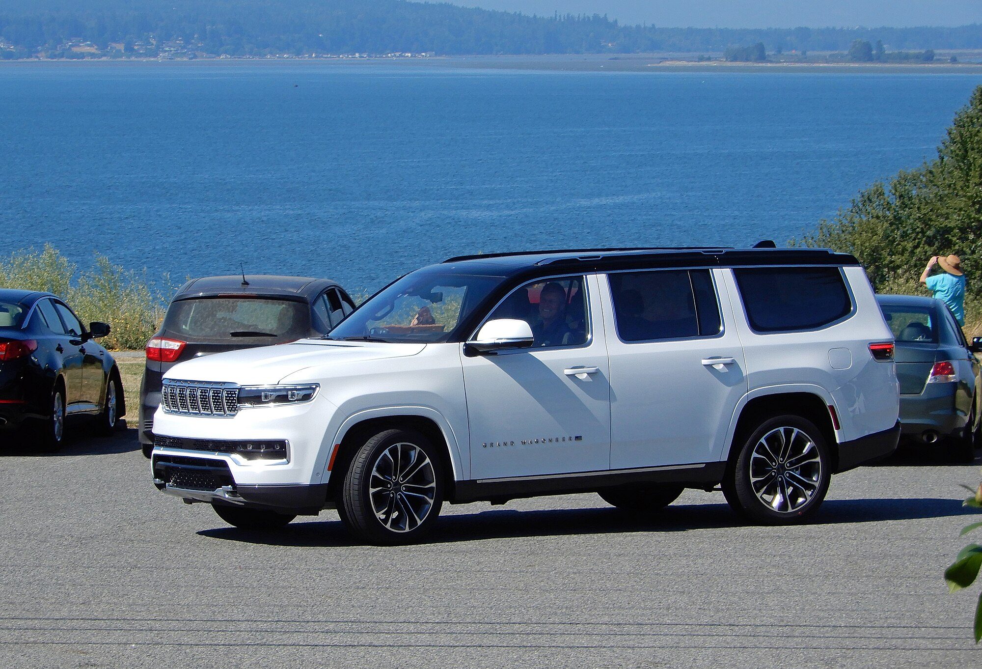A white 2022 Jeep Grand Wagoneer with a black roof at a paved waterfront pull-off above a wide blue bay