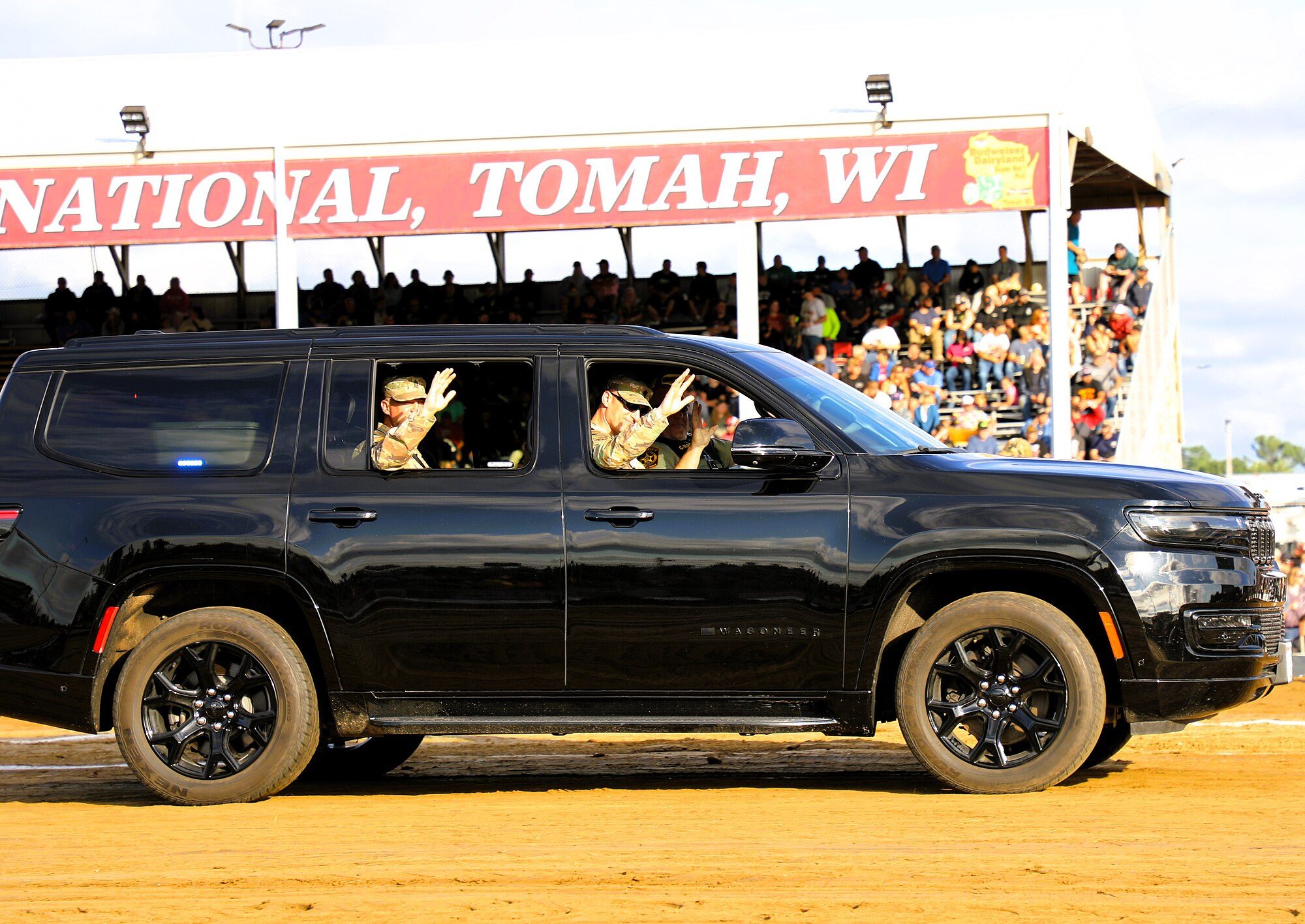 A black Jeep Wagoneer L with black wheels driving across the dirt arena of the Tomah, Wisconsin truck and tractor pull, with soldiers waving from its windows and a packed grandstand behind