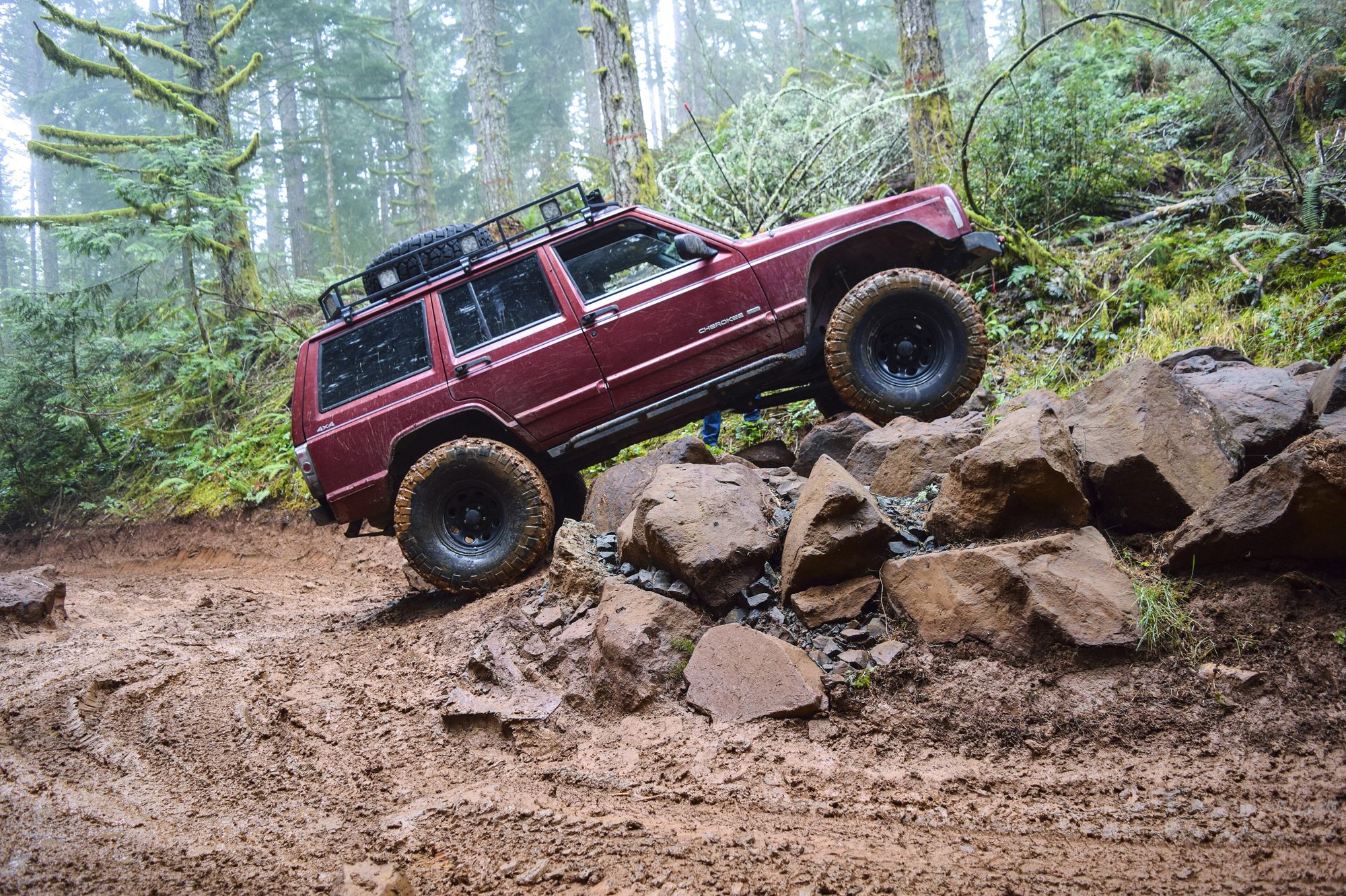 A lifted maroon 1997–2001 Jeep Cherokee XJ with a roof rack climbing a pile of boulders in a misty Oregon forest