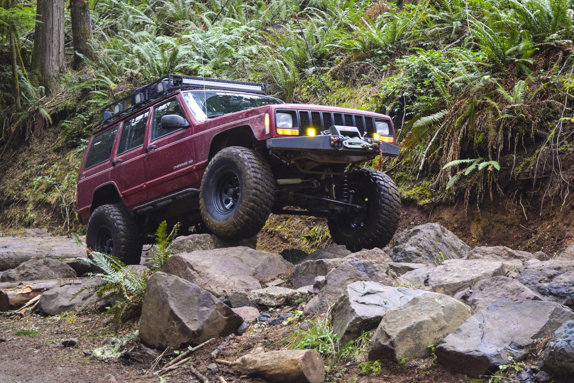 The same lifted maroon Cherokee XJ with a winch bumper crawling over rocks among ferns on a forest trail