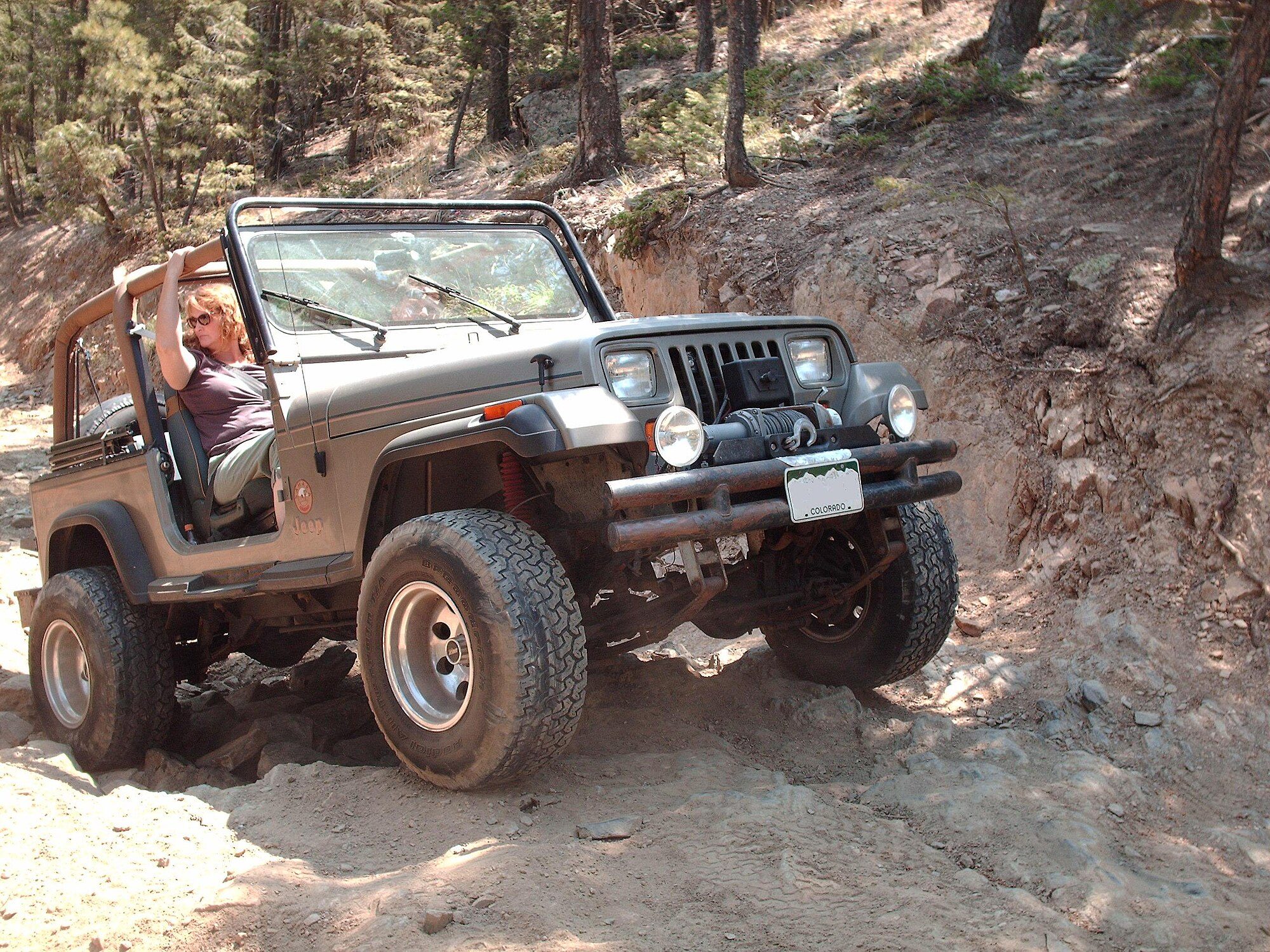 A grey-tan square-headlight Jeep Wrangler YJ with Colorado plates, no top and a winch bumper climbing a rocky dirt ledge in a pine forest