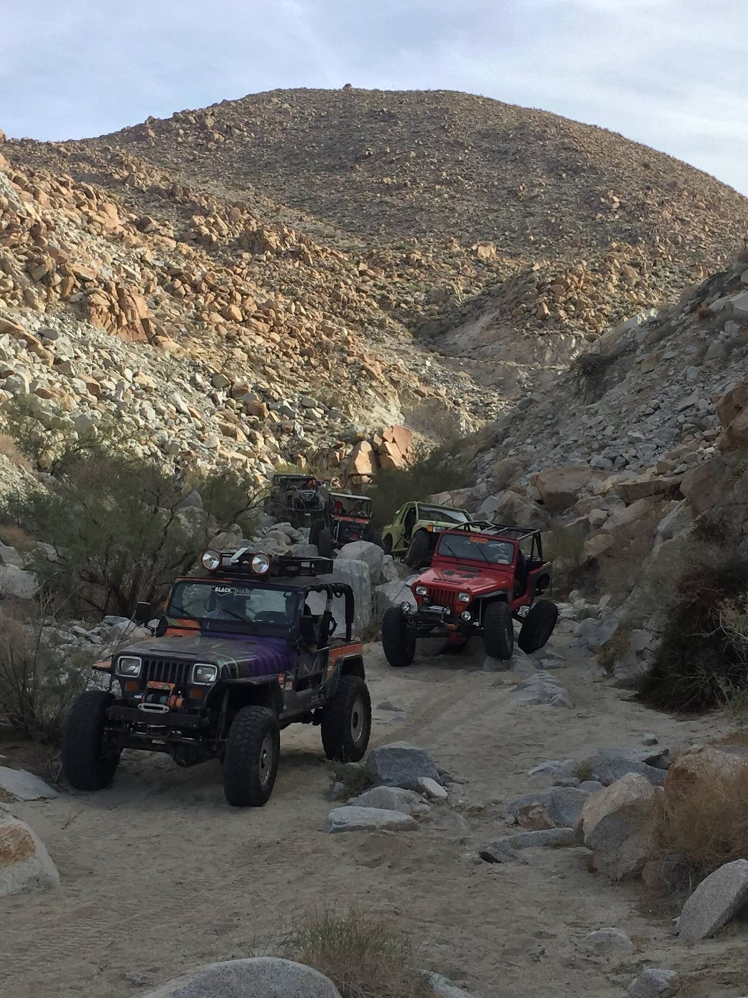 A lifted Jeep Wrangler YJ with roof lights leading a line of trail rigs up a rocky desert wash