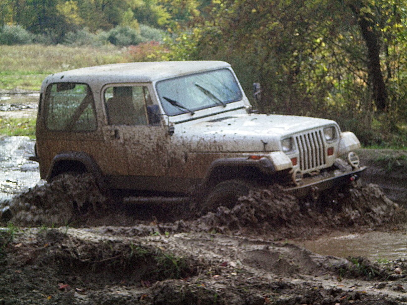 A white Jeep Wrangler YJ hardtop covered in mud, crossing a muddy creek bed