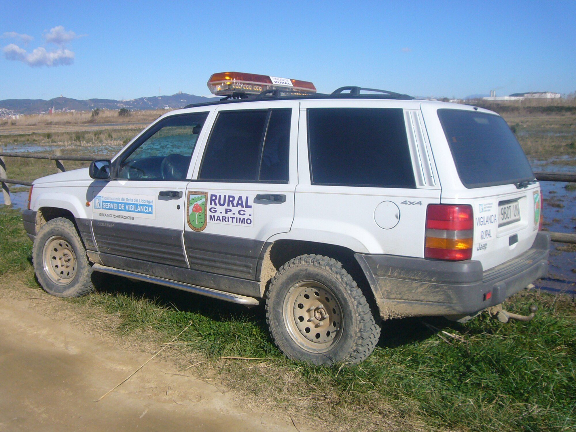 A white 1993–95 Grand Cherokee Laredo patrol truck with a light bar and muddy tires parked on grass beside wetlands