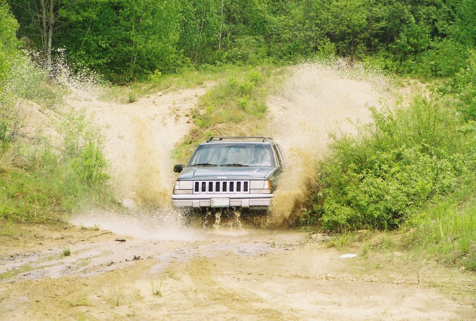 A grey-green 1994 Jeep Grand Cherokee ZJ fording a muddy puddle between green bushes, throwing up a wall of brown spray
