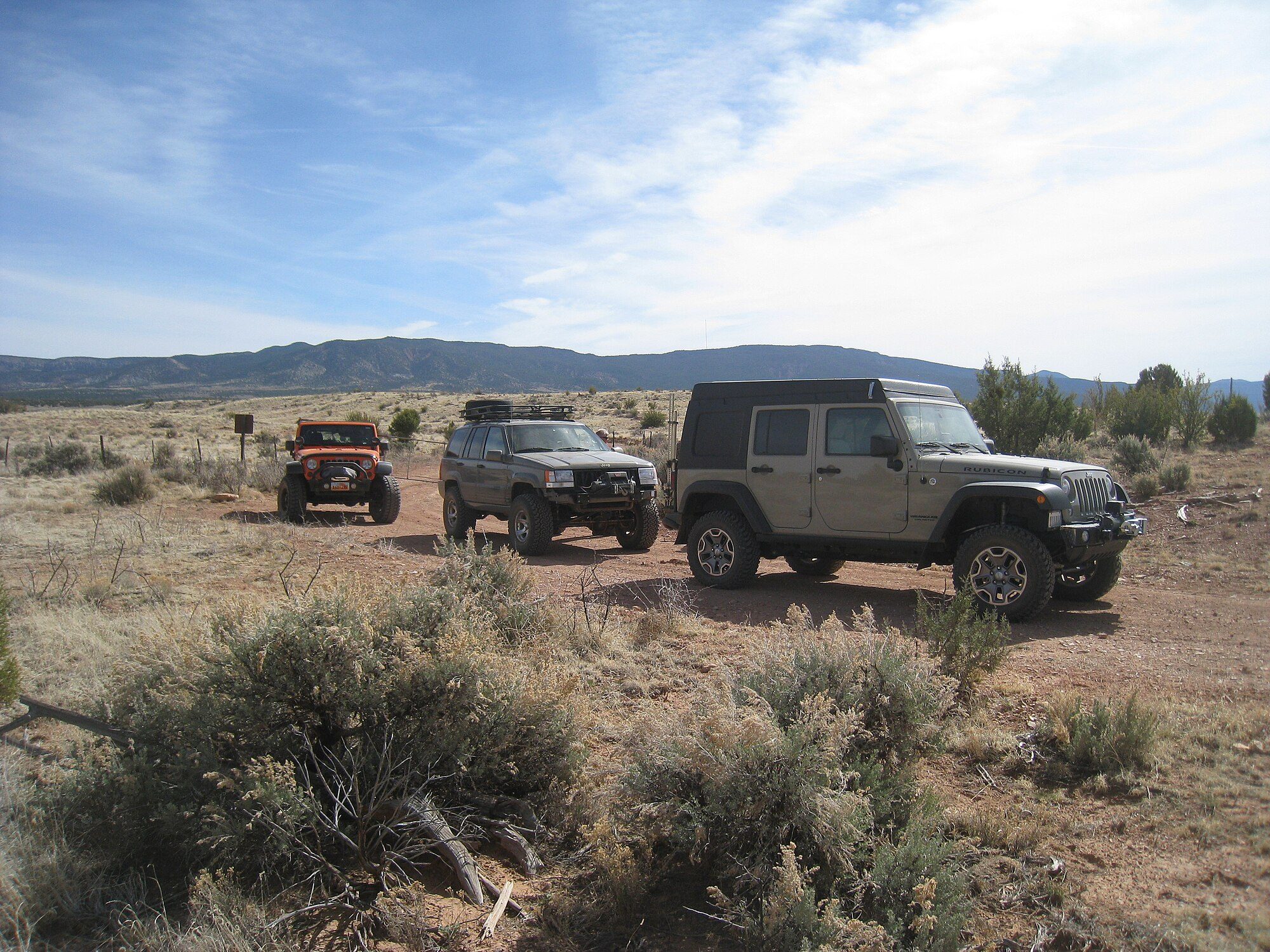 A lifted 1996–98 Grand Cherokee ZJ with a winch bumper and roof basket parked on a red dirt road in sagebrush country between two Wranglers