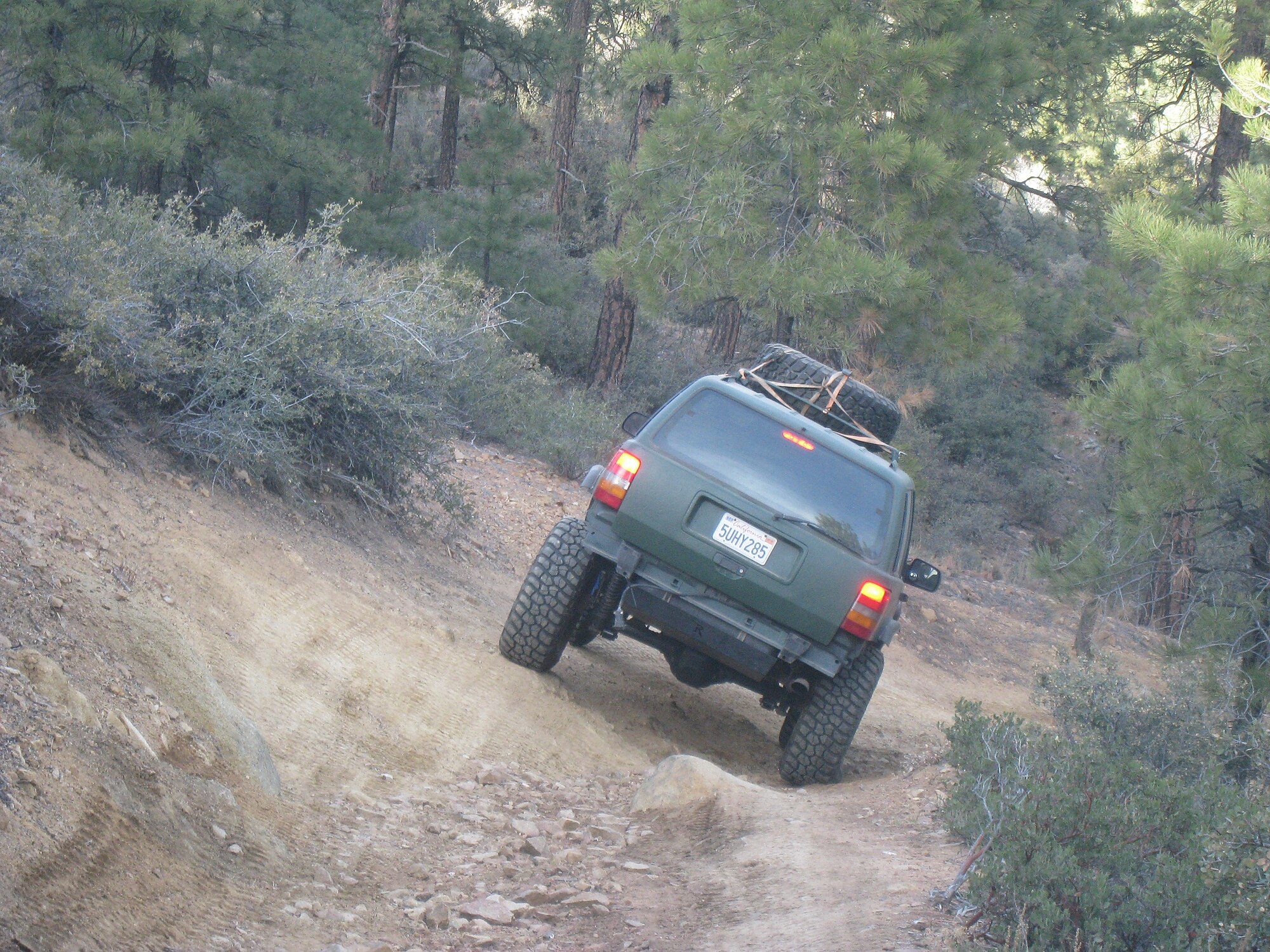 A lifted matte-green Grand Cherokee ZJ with a spare tire on the roof crawling up an off-camber dirt trail through pine forest