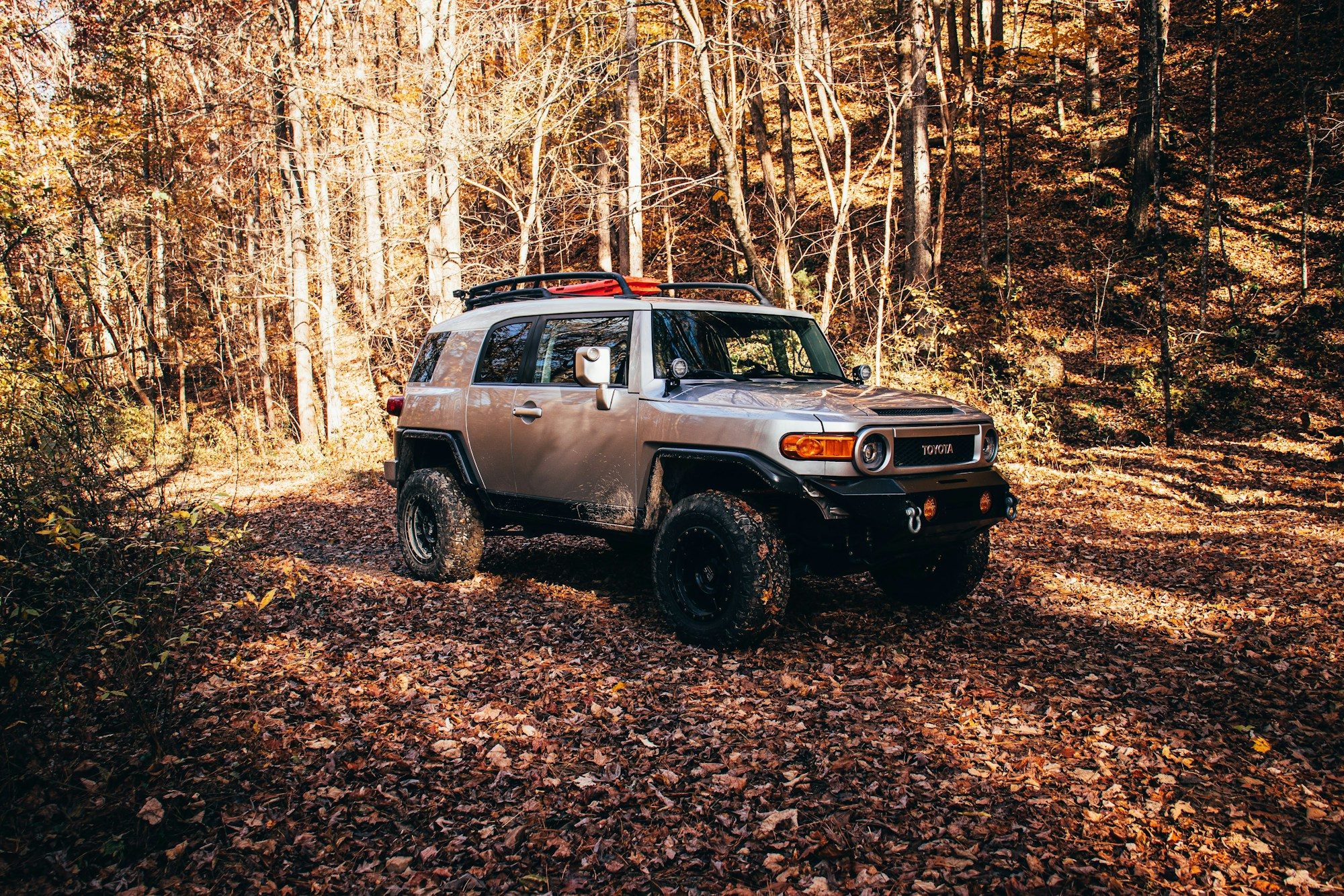 A silver FJ Cruiser with a roof rack and recovery boards on a leaf-covered forest trail in autumn
