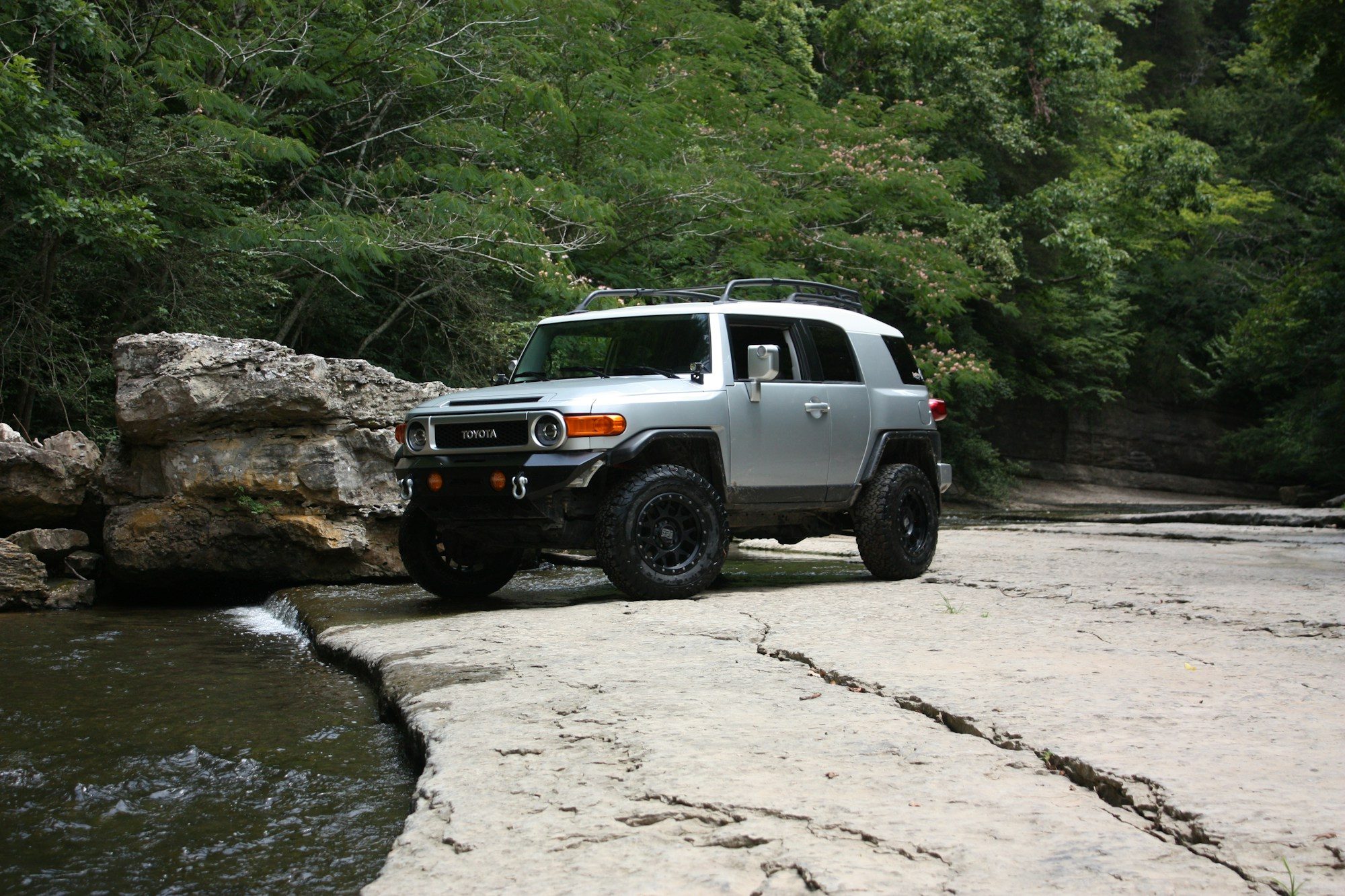 A lifted silver FJ Cruiser on all-terrain tires parked on flat creek-bed rock beside a small waterfall