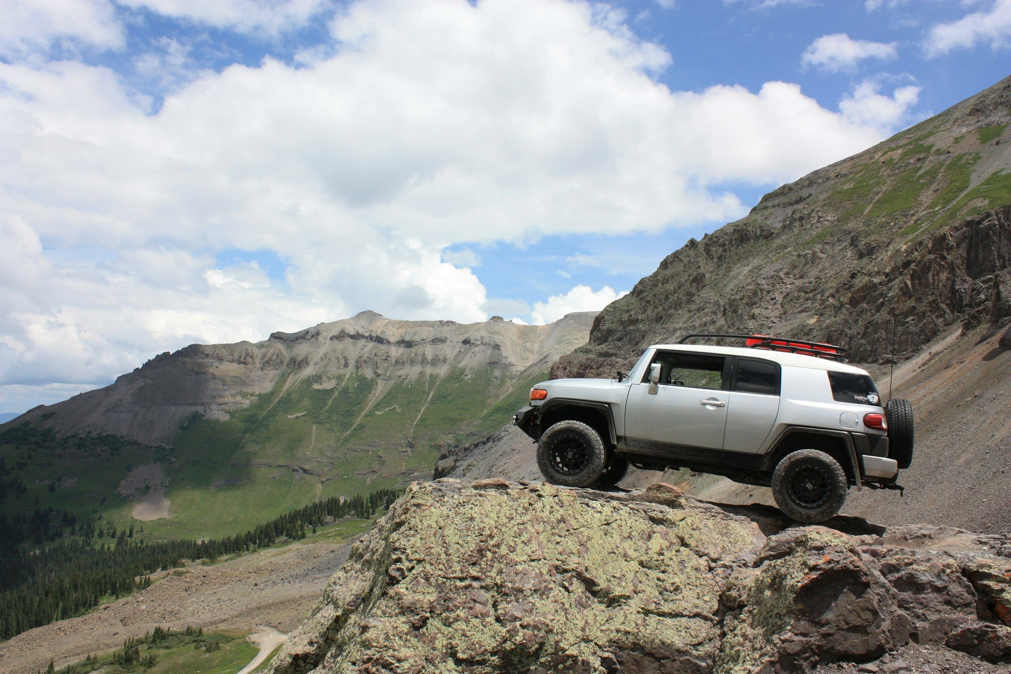A silver FJ Cruiser with a roof rack and black wheels perched on a lichen-covered rock ledge above a green alpine valley
