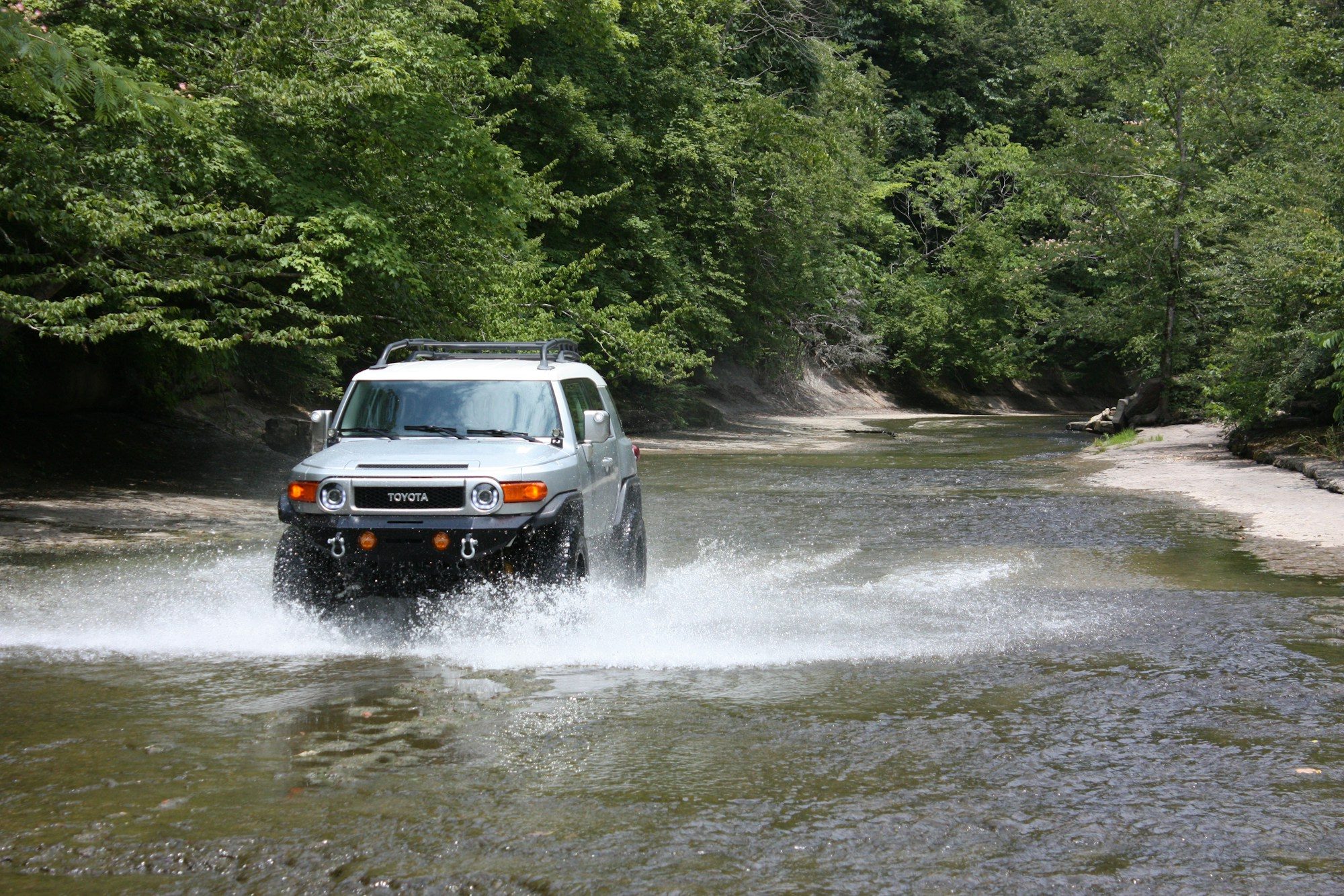 A silver FJ Cruiser with a steel bumper splashing through a shallow, tree-lined river