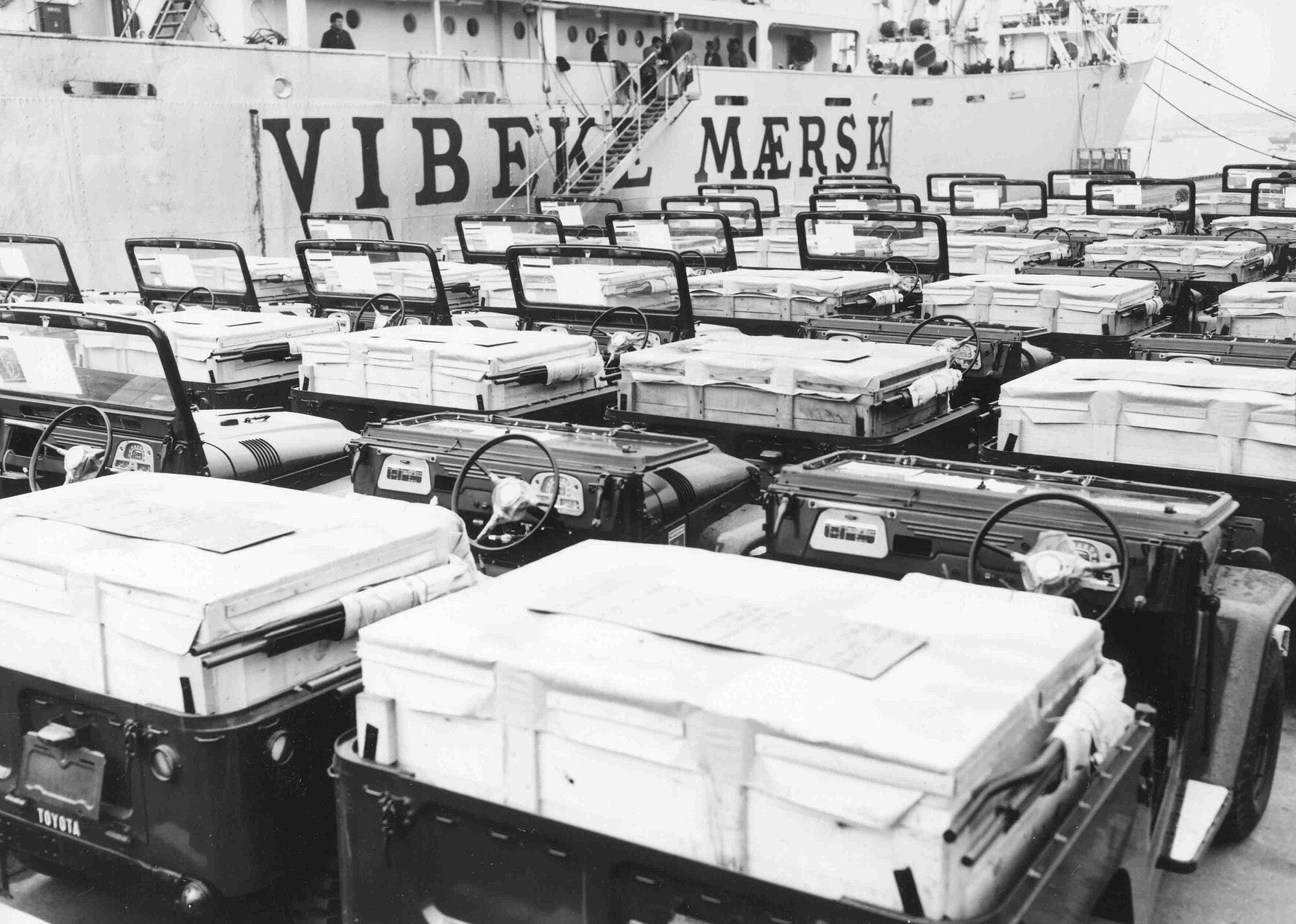 Black-and-white photo of rows of 20 Series Land Cruisers with crated loads in their rear tubs, lined up on a quay beside the Maersk Line ship Vibeke Maersk