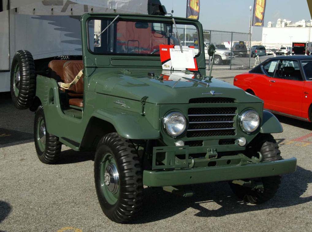 A restored olive-green FJ25 Land Cruiser with a fold-down windshield, side-mounted spare and round headlights, parked at an outdoor Toyota show