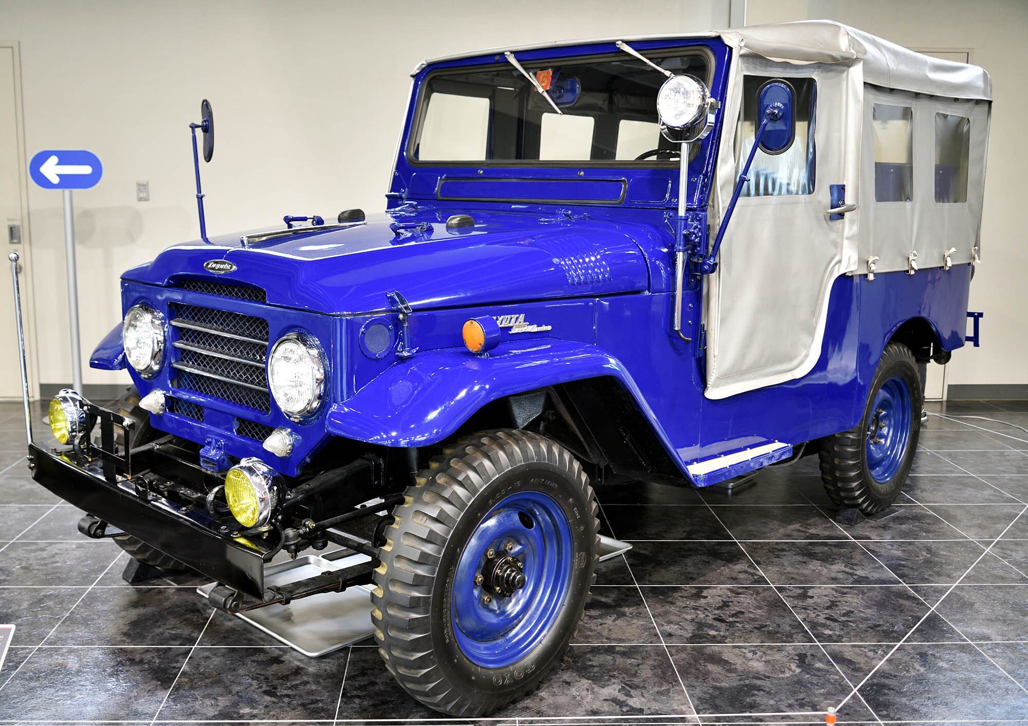 A blue 1957 FJ25L Land Cruiser with a white canvas top and blue steel wheels on display indoors