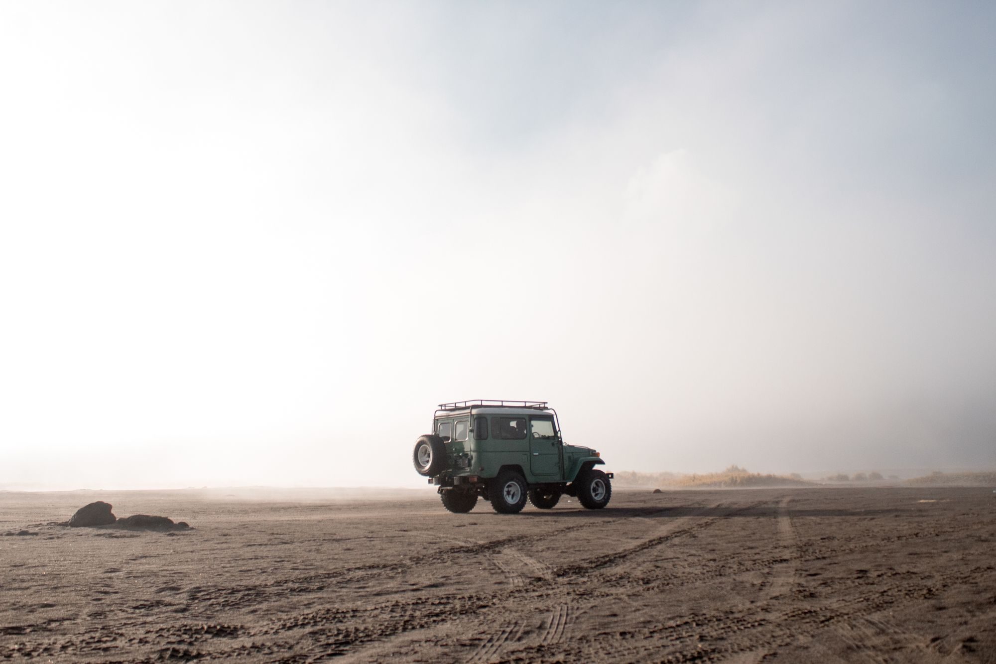A green short-wheelbase FJ40 hardtop with a roof rack and rear-mounted spare crossing the volcanic sand sea below Mount Bromo in morning mist