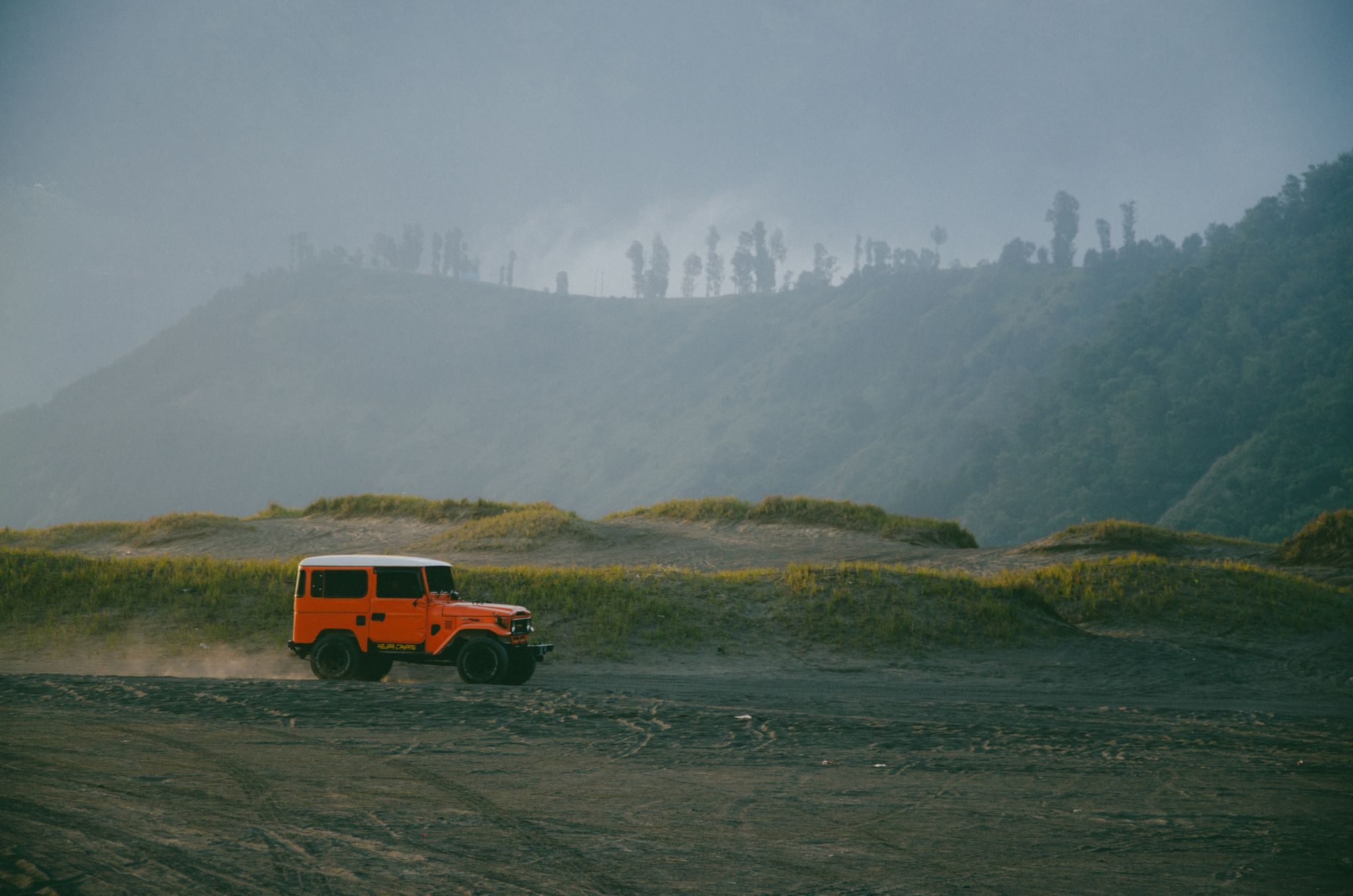 An orange short-wheelbase FJ40 hardtop with a white roof kicking up dust on a black-sand track between grassy dunes at Bromo