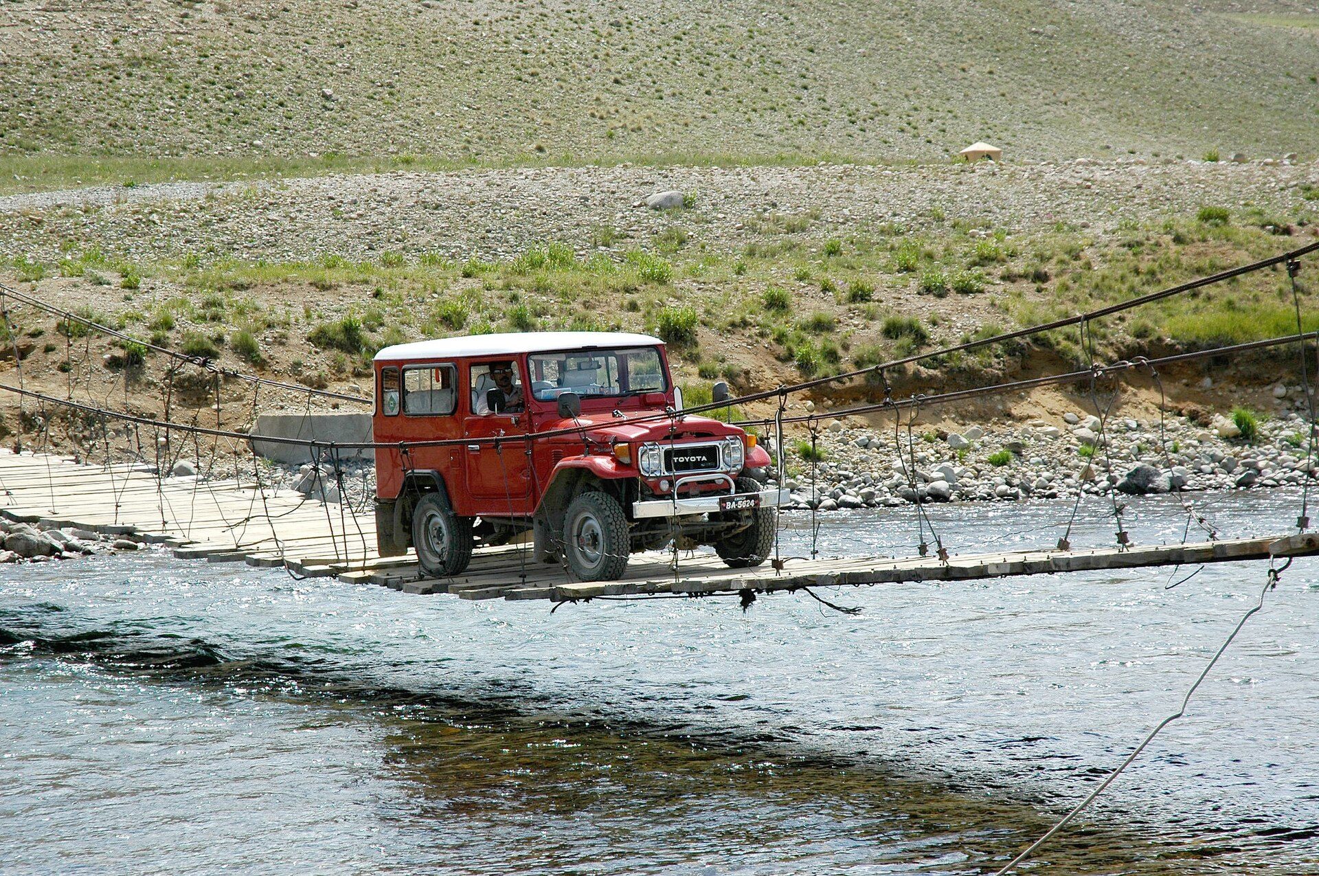 A red 40 Series Land Cruiser hardtop with a white roof and square headlight bezels crossing a timber-and-cable suspension bridge over a river on the Deosai plateau