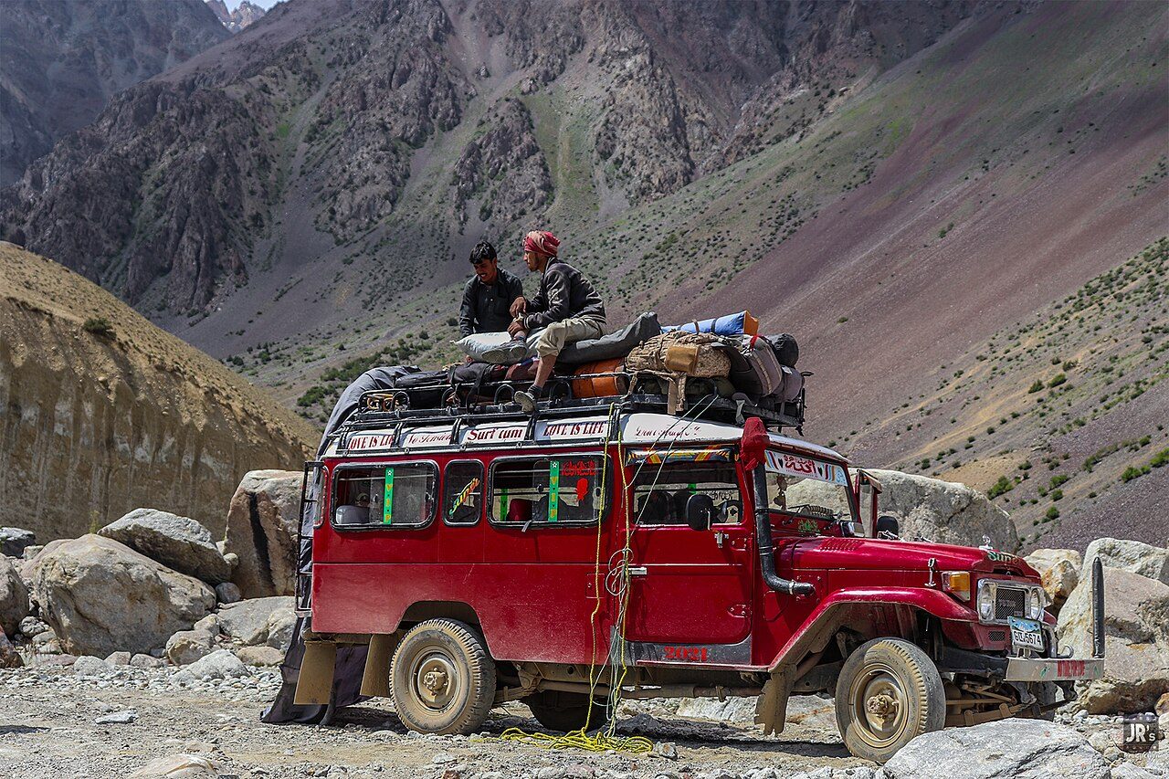 A red long-wheelbase 40 Series Land Cruiser hardtop with square headlight bezels, loaded with passengers and luggage on its roof, on a boulder-strewn mountain track