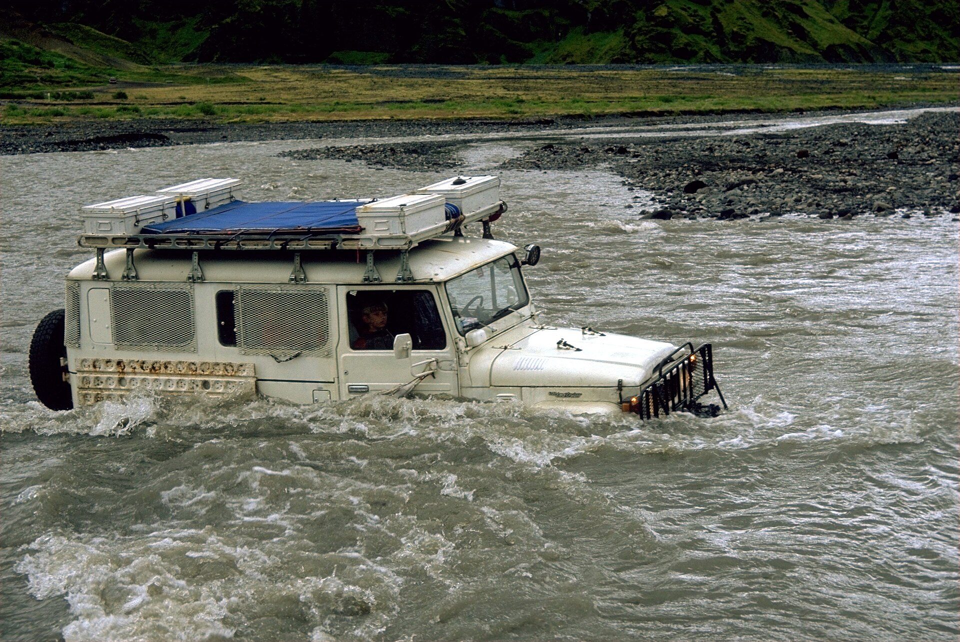 A long-wheelbase Land Cruiser 40 Series camper fording a fast glacial river up to its bumper