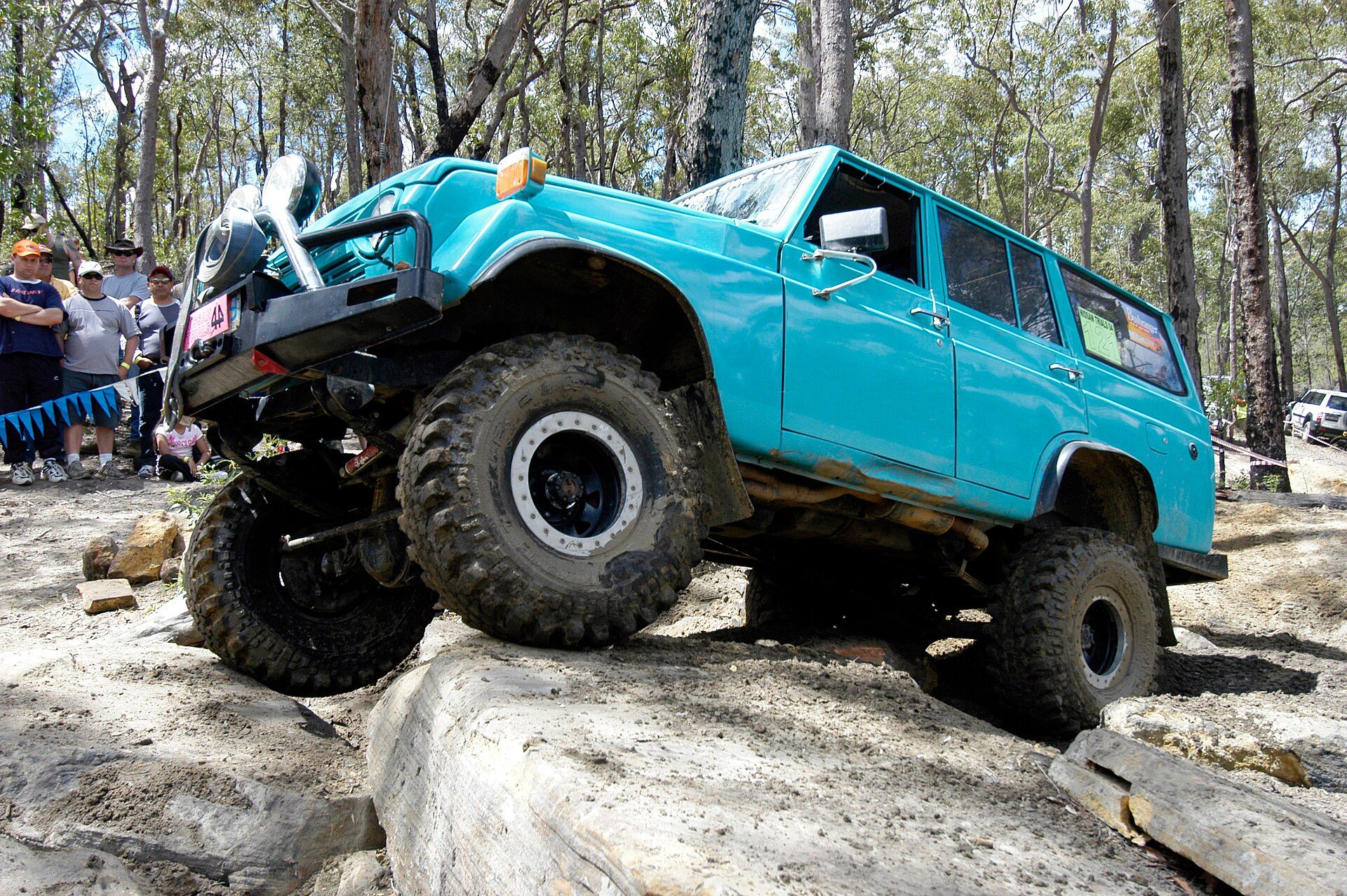 A lifted turquoise Land Cruiser FJ55 wagon flexing over a rock step at a 4WD trials event