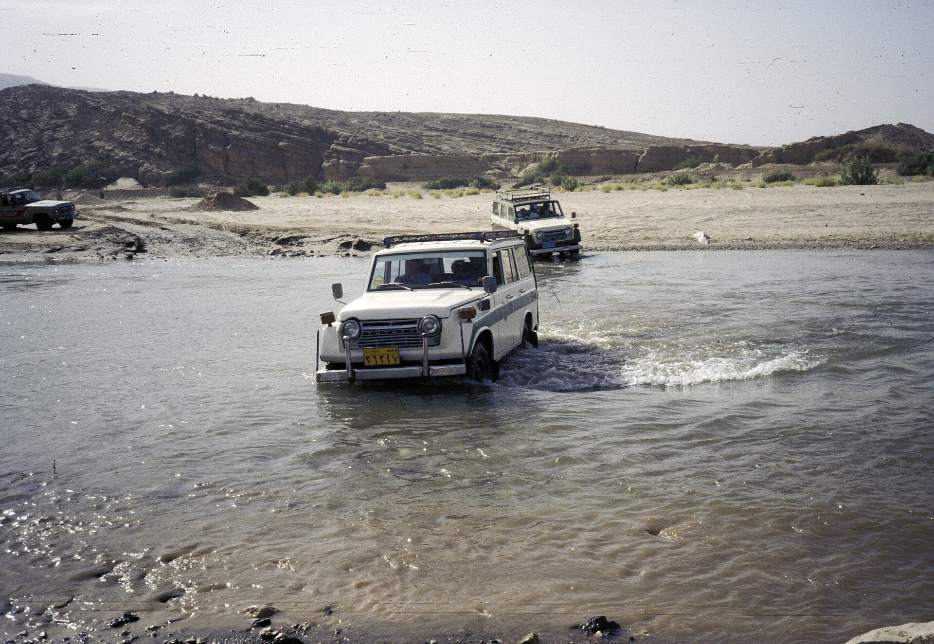 A white Land Cruiser FJ55 wagon with a roof rack fording a shallow river in a Yemeni wadi, with a 60 Series following behind