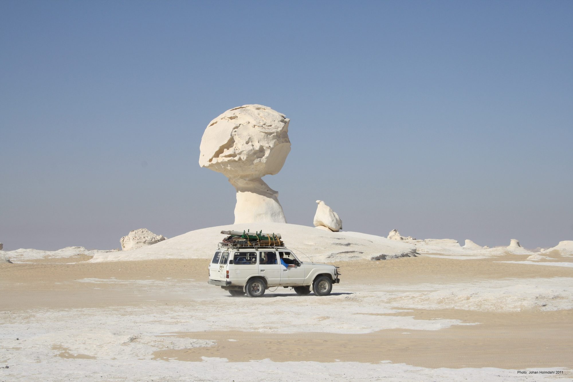 A white Land Cruiser 60 Series wagon with a loaded roof rack driving past a mushroom-shaped chalk formation in Egypt's White Desert