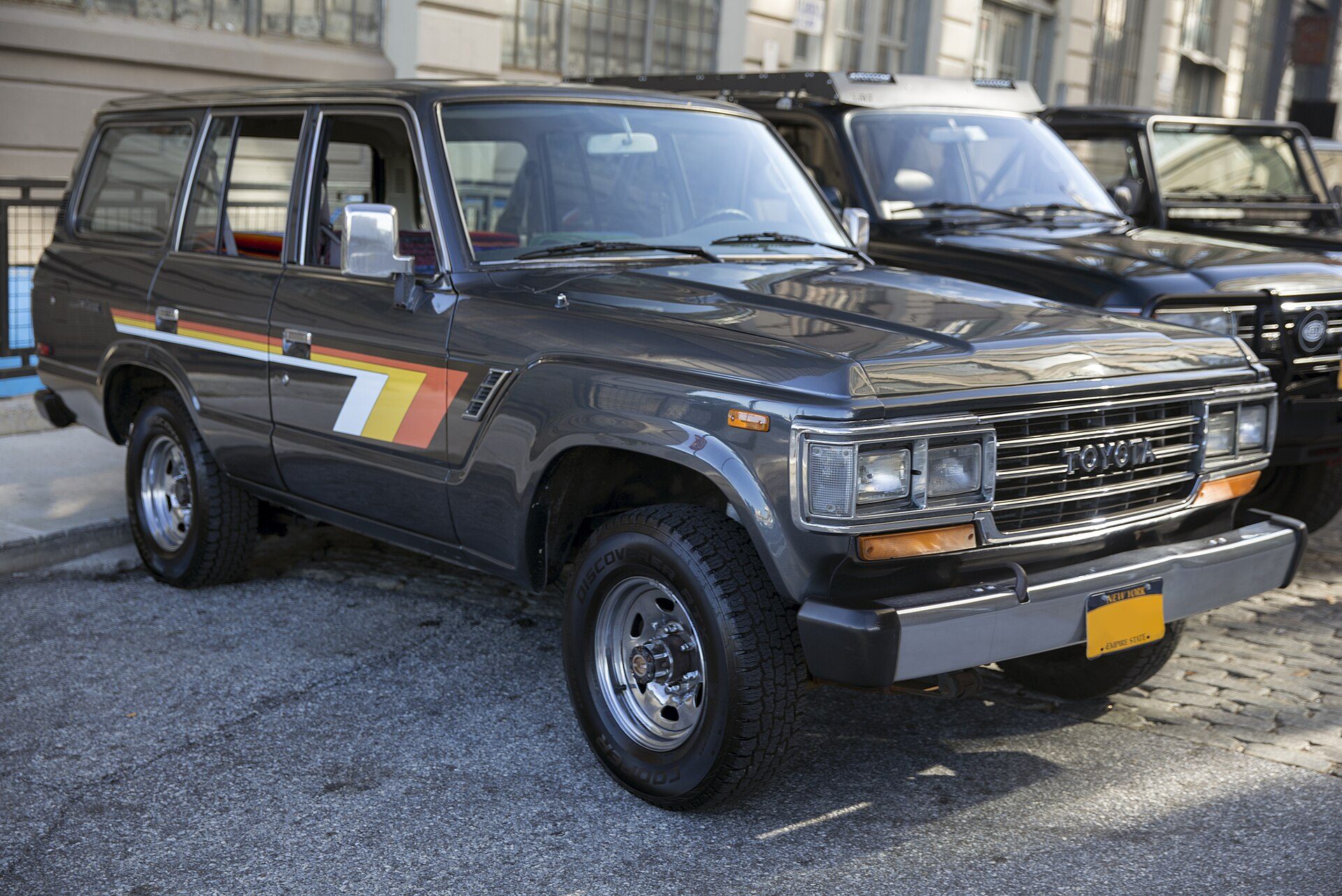 A grey metallic 1990 US-market Land Cruiser FJ62 with four rectangular headlights and rainbow side stripes, parked on a street