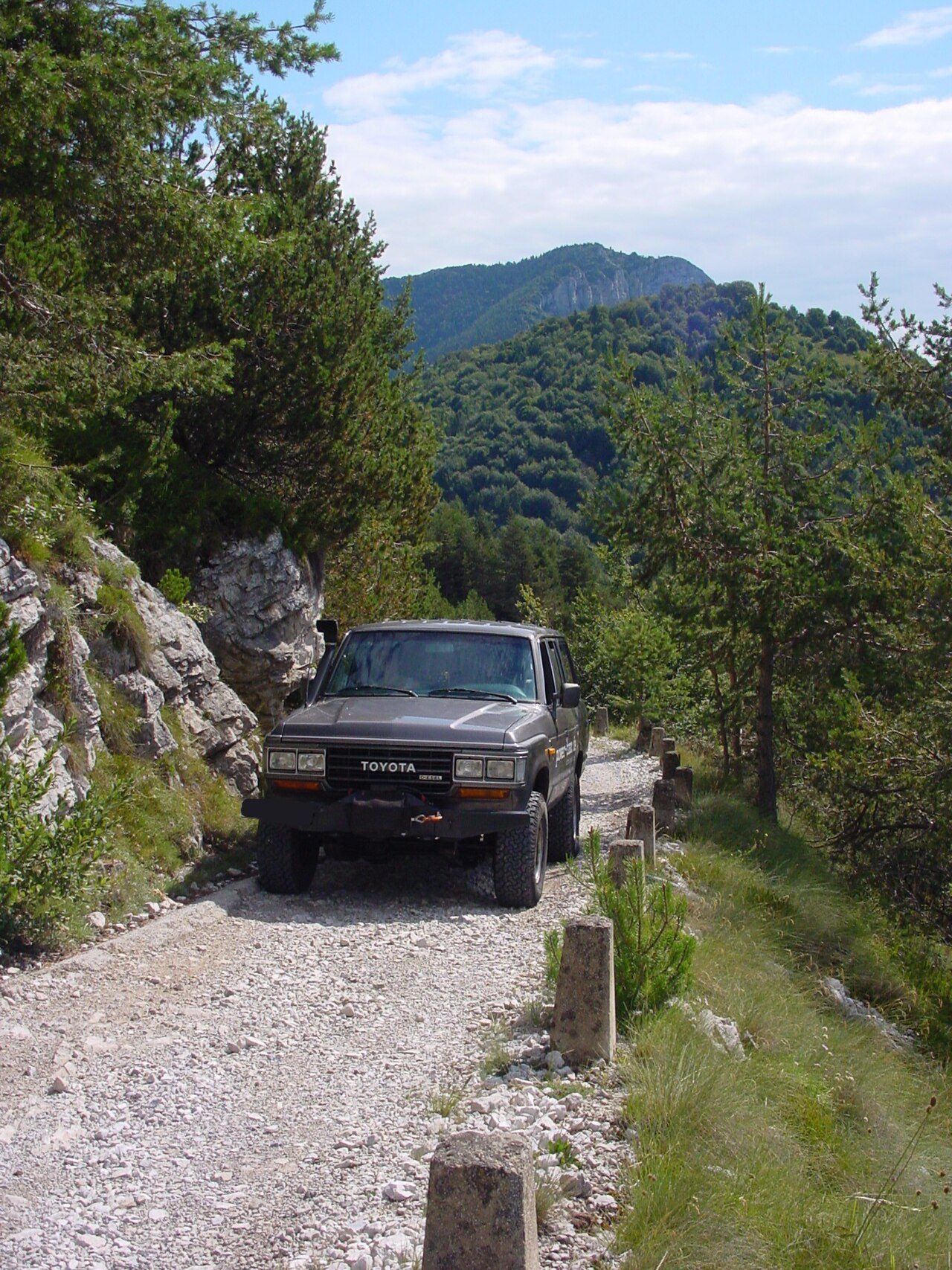 A dark late 60 Series Land Cruiser diesel with the four-headlight front, a winch bumper and a snorkel on a narrow gravel mountain road at Passo di Tremalzo