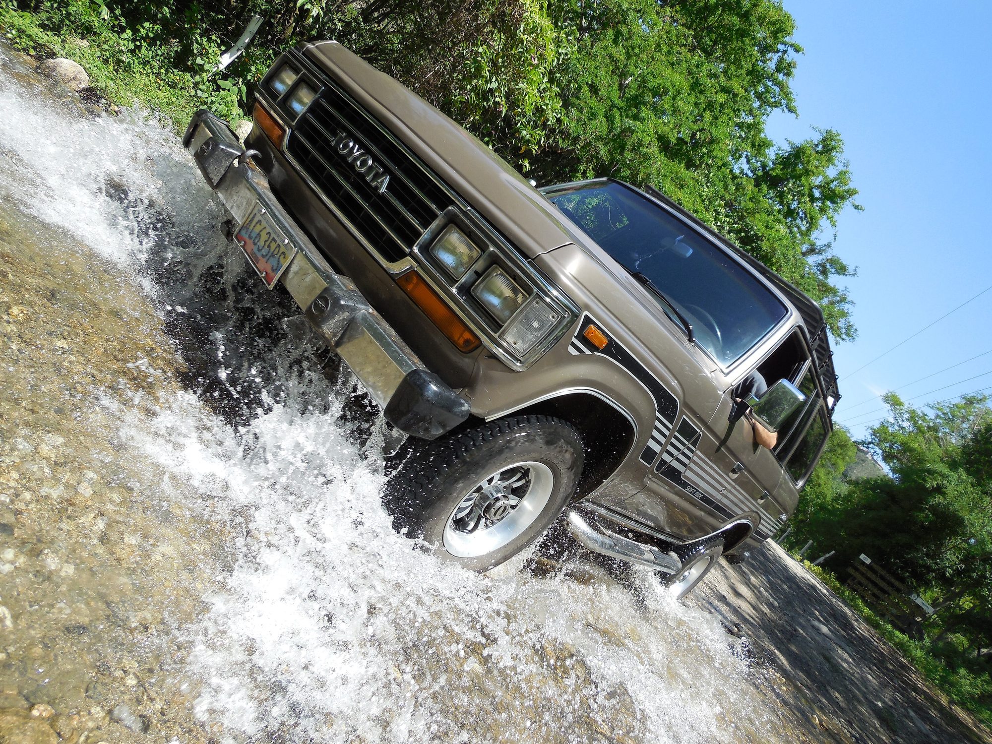 A bronze 1992 Venezuelan-market Land Cruiser FJ62 GX with four rectangular headlights splashing through a rocky stream crossing, shot from a low tilted angle