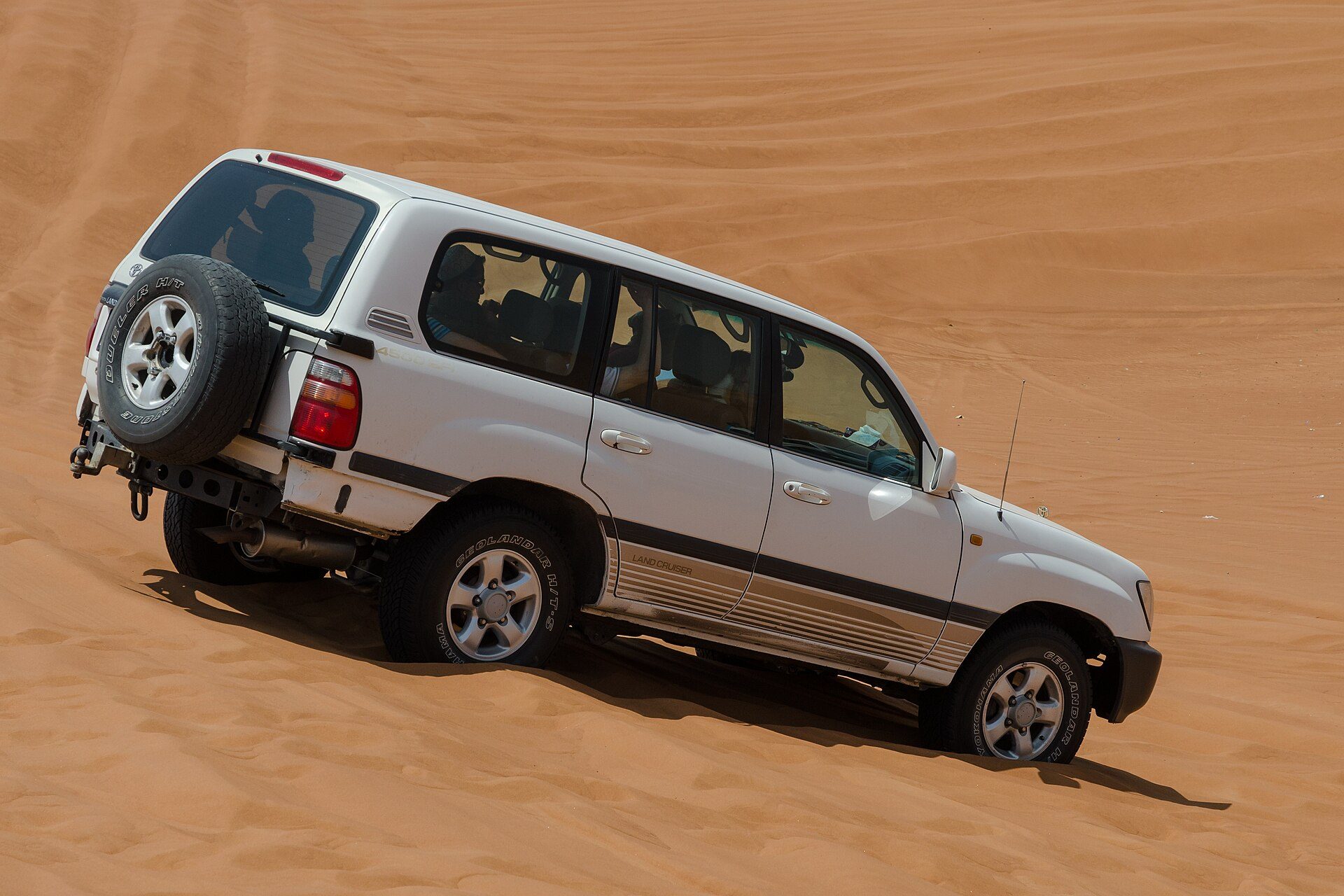 A white Land Cruiser 100 Series with a 4500 EFI badge and rear-mounted spare wheel side-hilling across a sand slope in the UAE