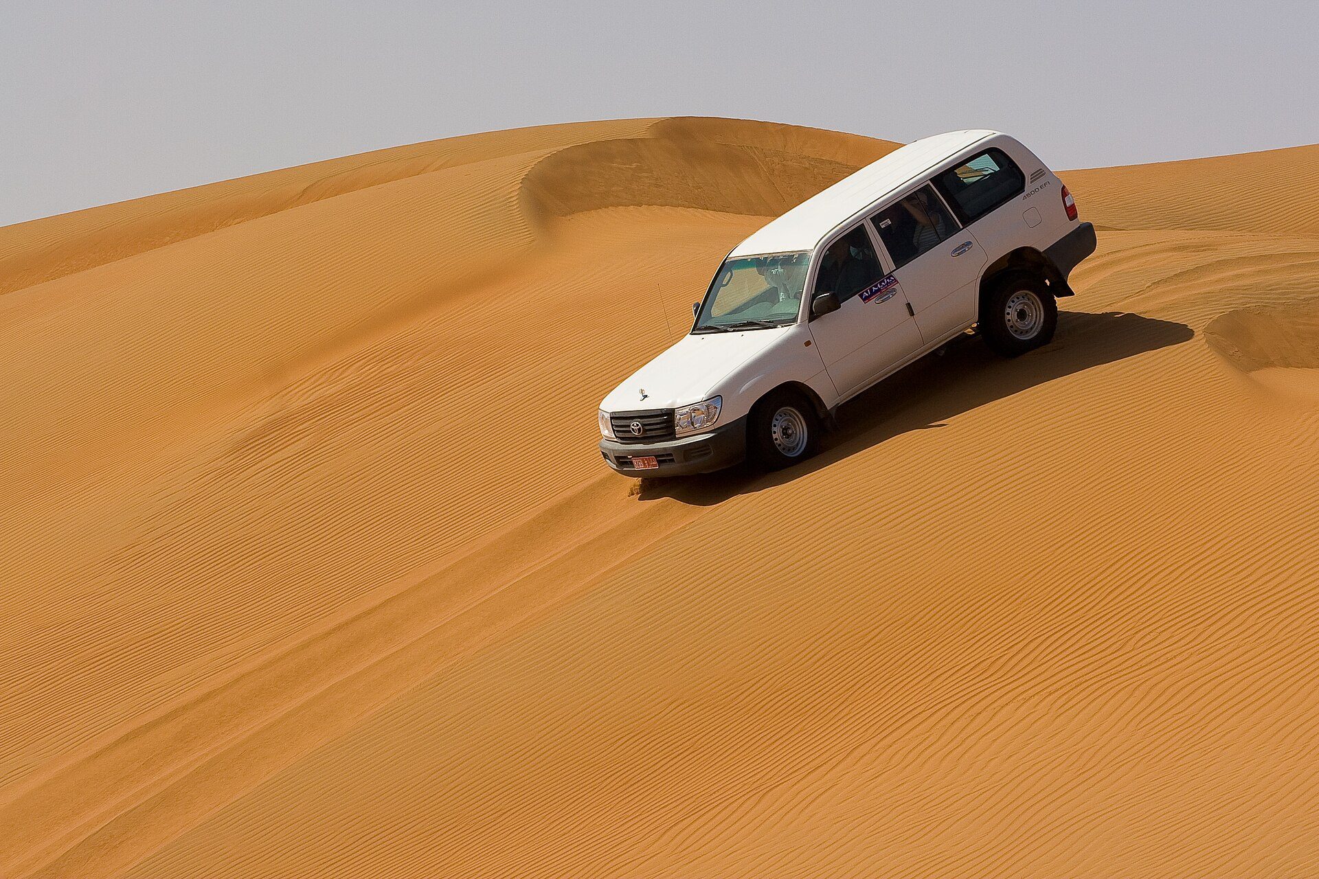 A white, steel-wheeled Land Cruiser 100 Series with a 4500 EFI badge running along the face of a tall dune in the Wahiba Sands, Oman