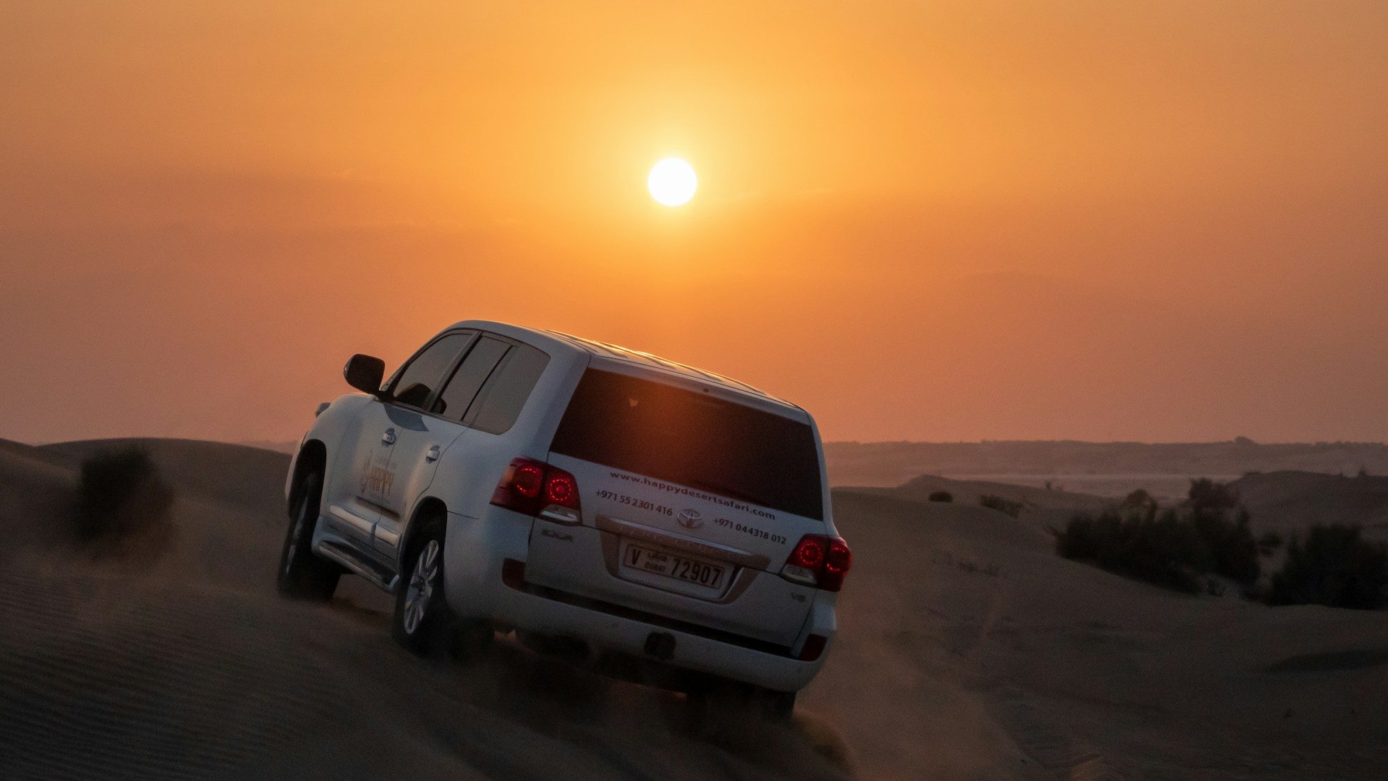 A white facelifted Land Cruiser 200 Series safari truck climbing a sand dune near Dubai at sunset